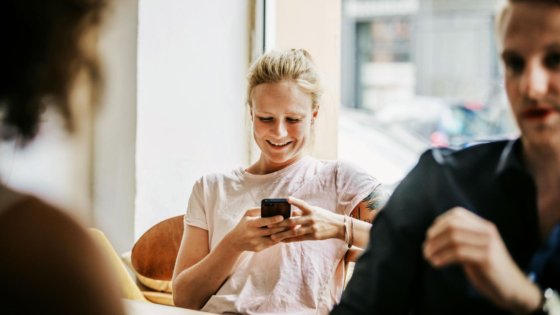 Lächelnde junge Frau sitzt in Café und schaut auf ihr Smartphone.