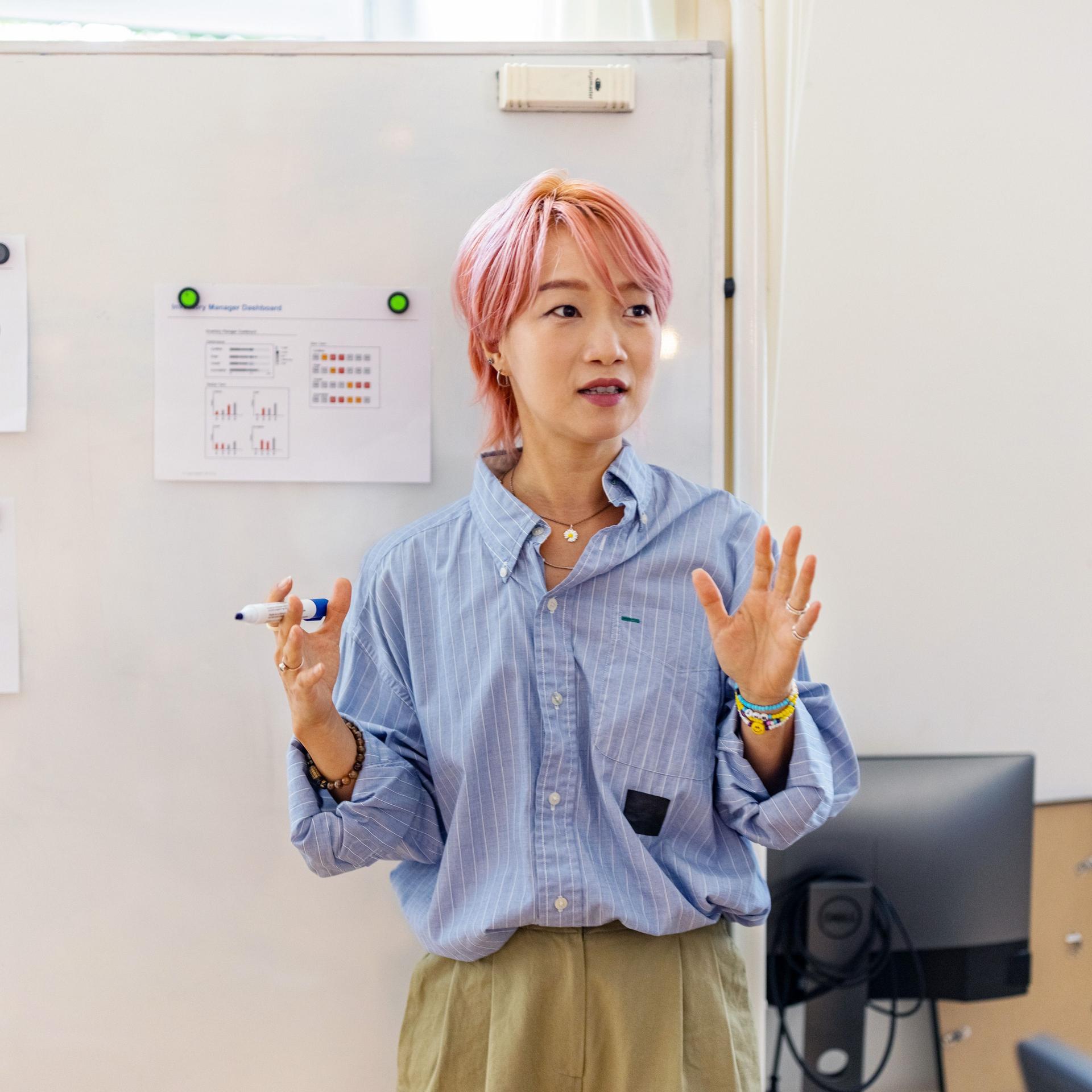 Junge Frau mit rosa gefärbten Haaren steht gestikulierend vor einem Whiteboard mit Charts und Graphen