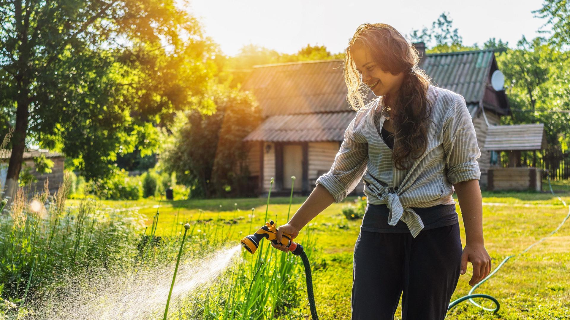 Junge Frau bewässert mit einem Gartenschlauch Pflanzen im Garten. Im Hintergrund steht ein Landhaus.