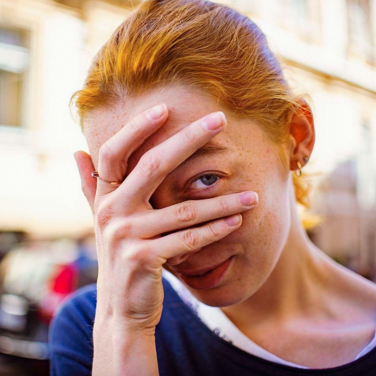 Straßenportrait einer jungen Frau, die ihr Gesicht mit ihrer Hand bedeckt. Sie schaut mit ihrem linken Auge zwischen Ring- und Mittelfinger hindurch.