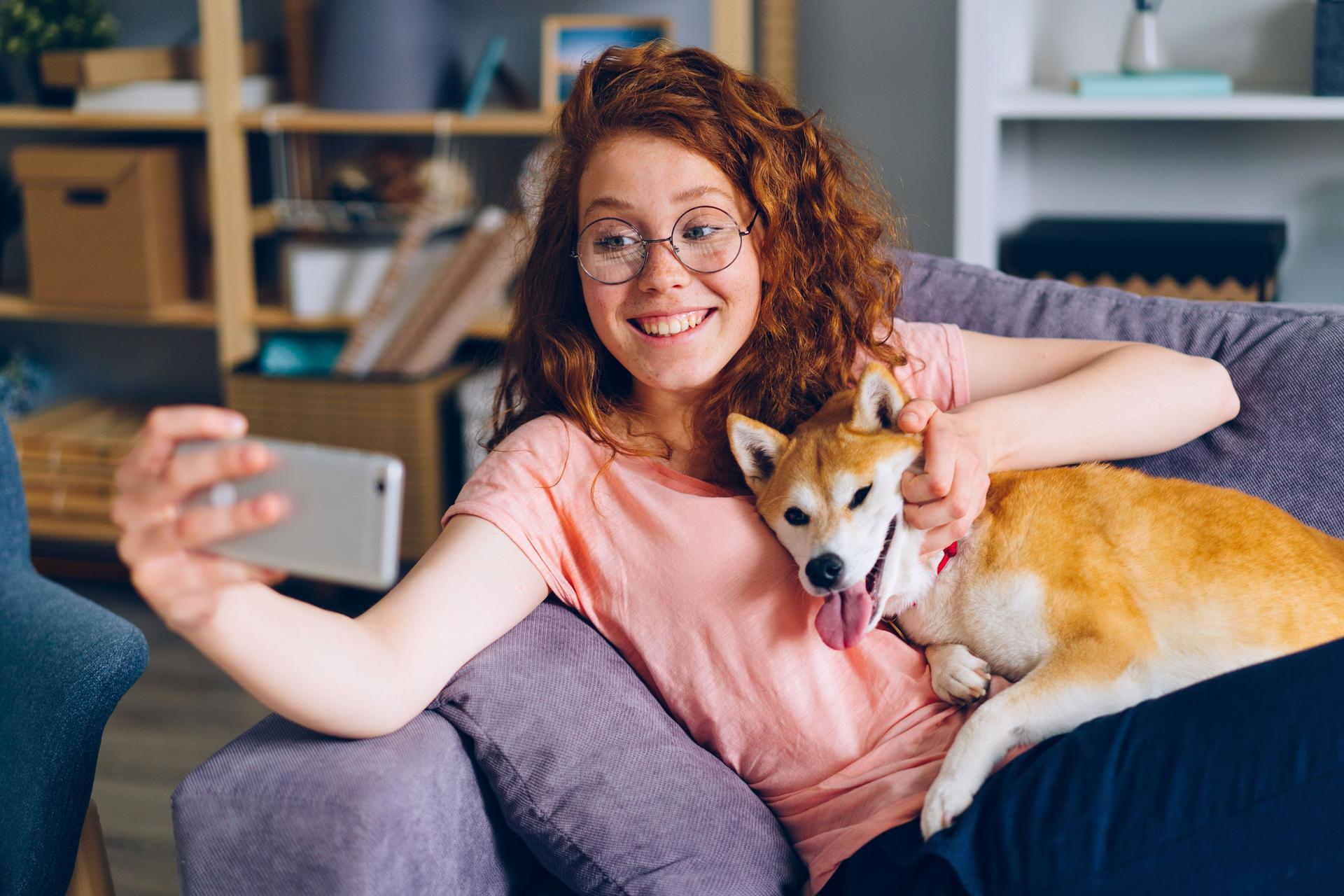 Junge Frau sitzt mit einem Shiba Inu Hund auf einem Sofa und macht mit einem Smartphone ein Selfie von sich und dem Hund.