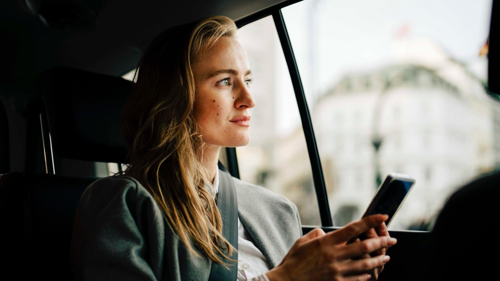 Nachdenkliche junge Frau mit Smartphone in der Hand. Sie sitzt auf der Rückbank und schaut durch das Autofenster in die Landschaft.