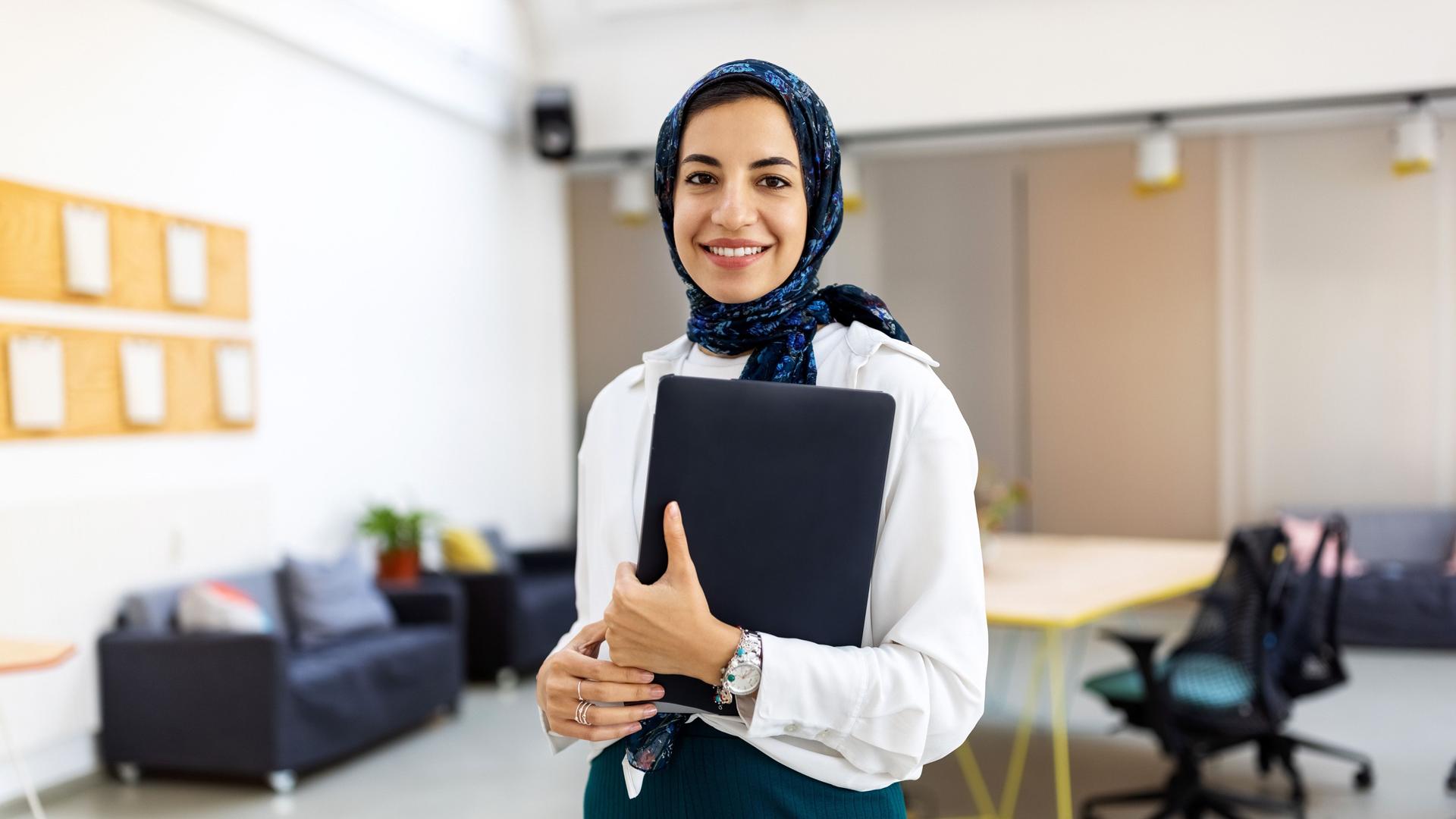 Eine junge Frau mit Hijab steht in einem großen Büro. Sie hält Unterlagen in einem Ordner in der Hand.