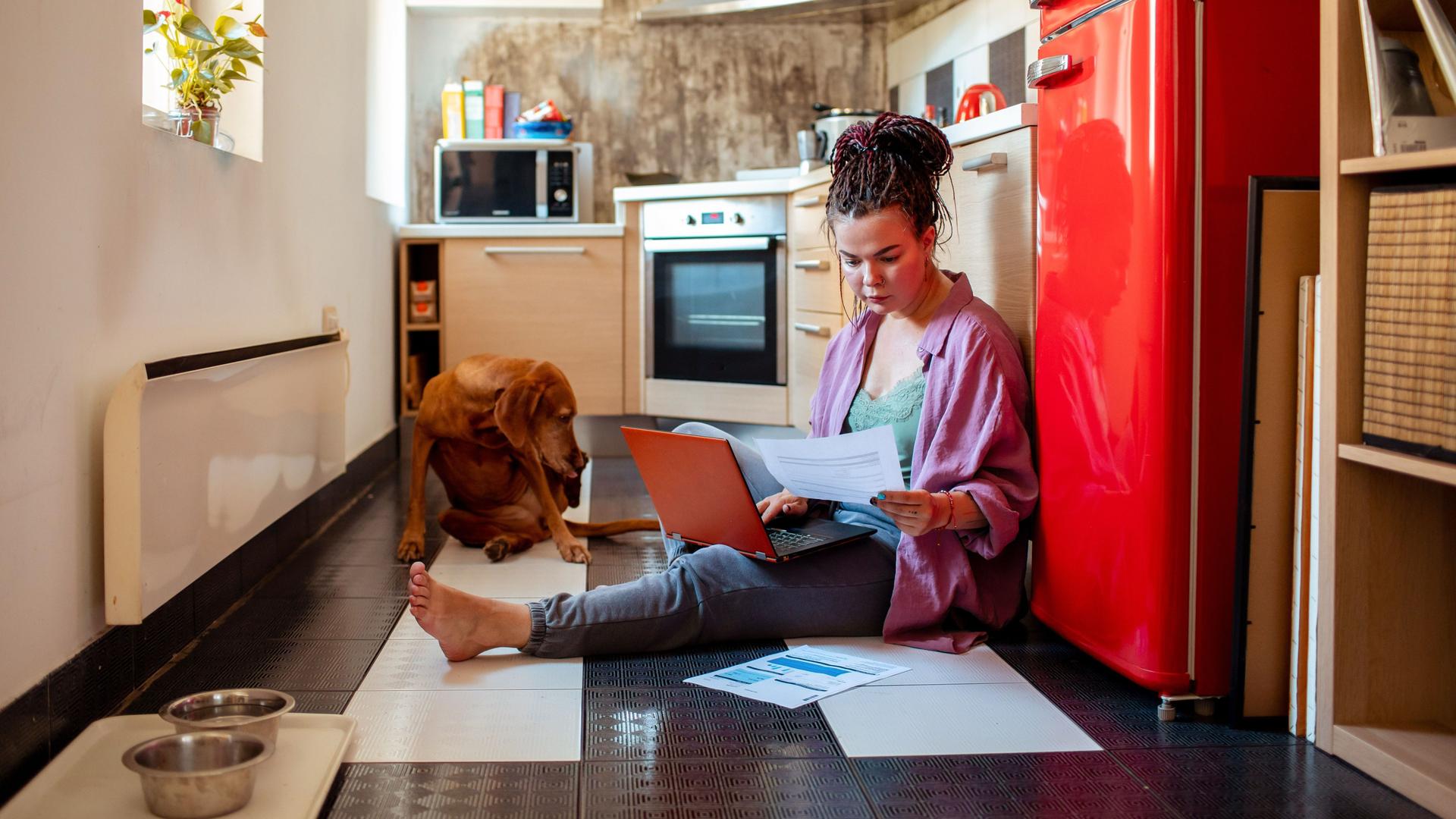Junge Frau sitzt mit rotem Laptop und Papierdokumenten vor einem roten Kühlschrank auf dem Küchenboden. Neben ihr liegt ein brauner Hund.