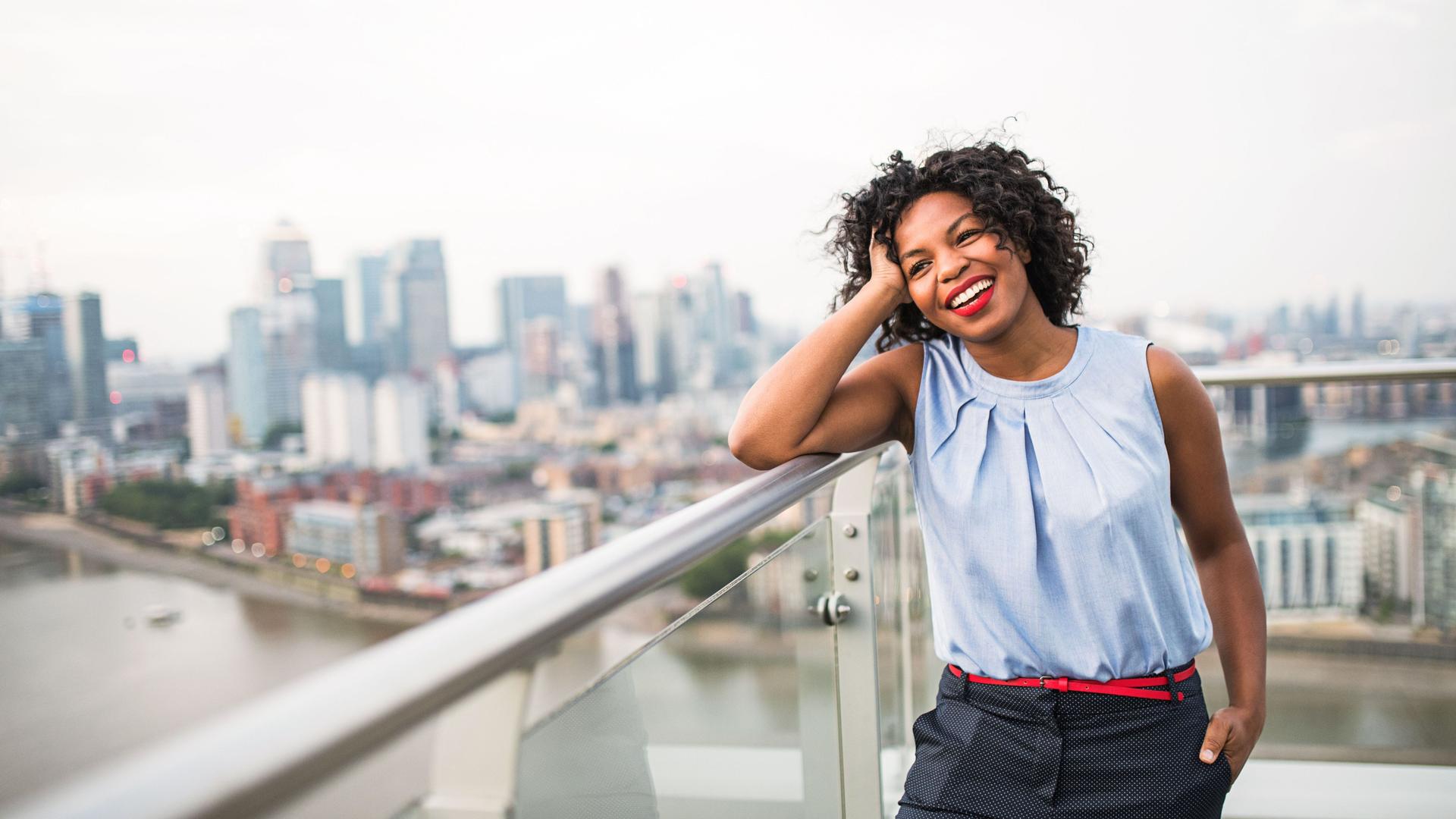 Junge Frau lehnt lachend auf Brückengeländer mit Großstadtkulisse im Hintergrund.