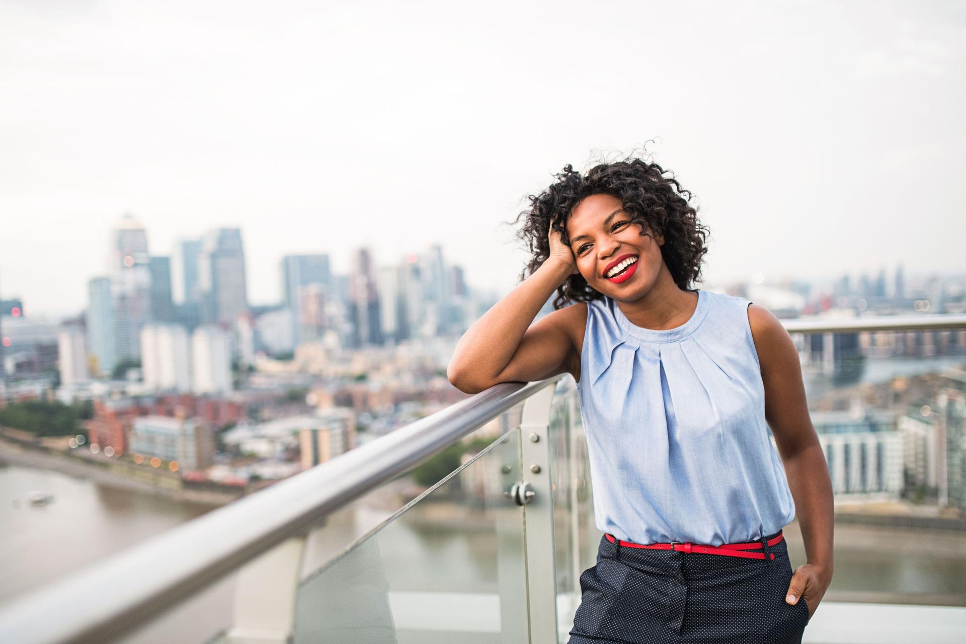 Junge Frau lehnt lachend auf Brückengeländer mit Großstadtkulisse im Hintergrund.