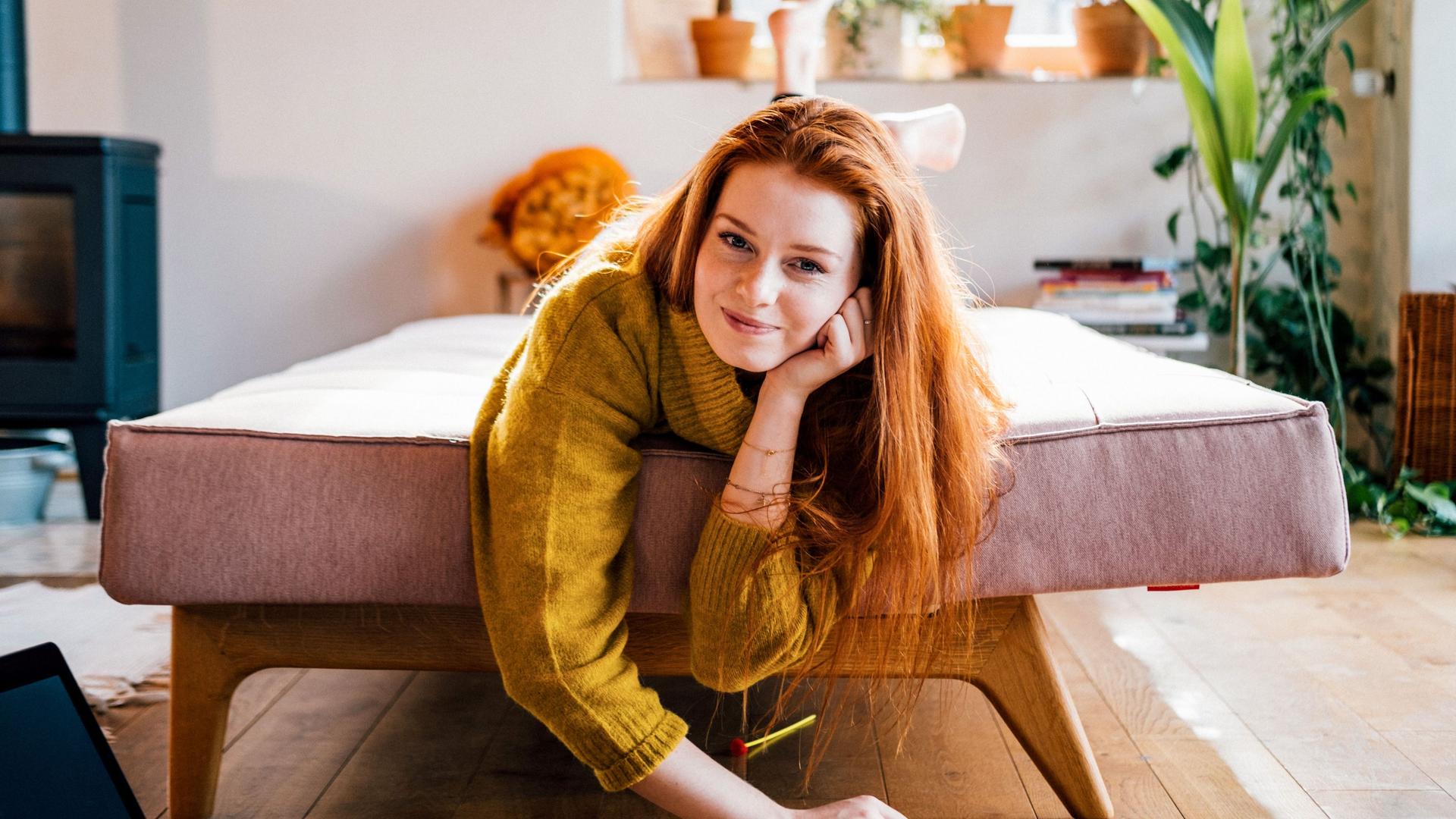 Portrait einer jungen Frau, die lächelnd im Wohnzimmer auf einem ausgezogenen Sofa liegt.