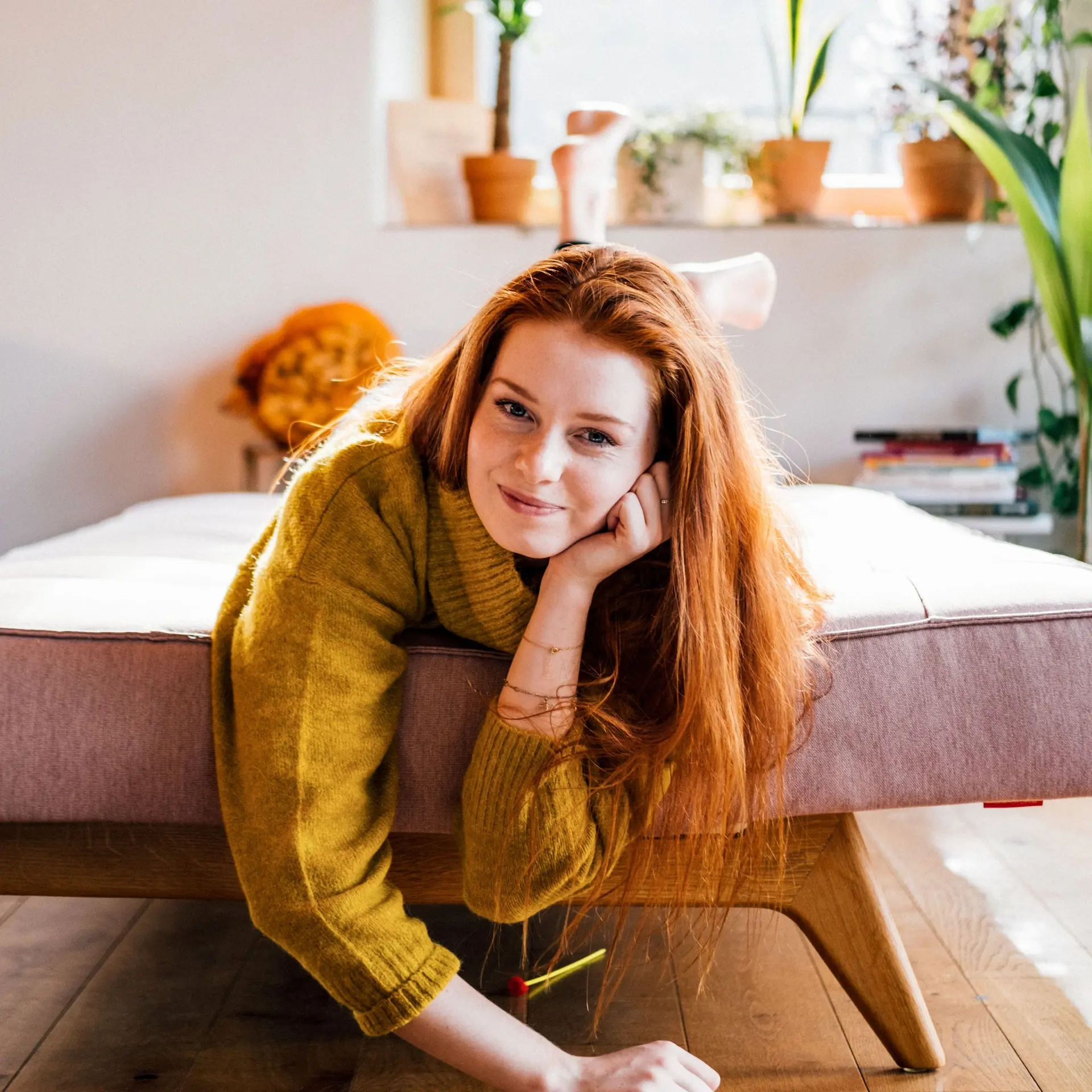 Portrait einer jungen Frau, die lächelnd im Wohnzimmer auf einem ausgezogenen Sofa liegt.