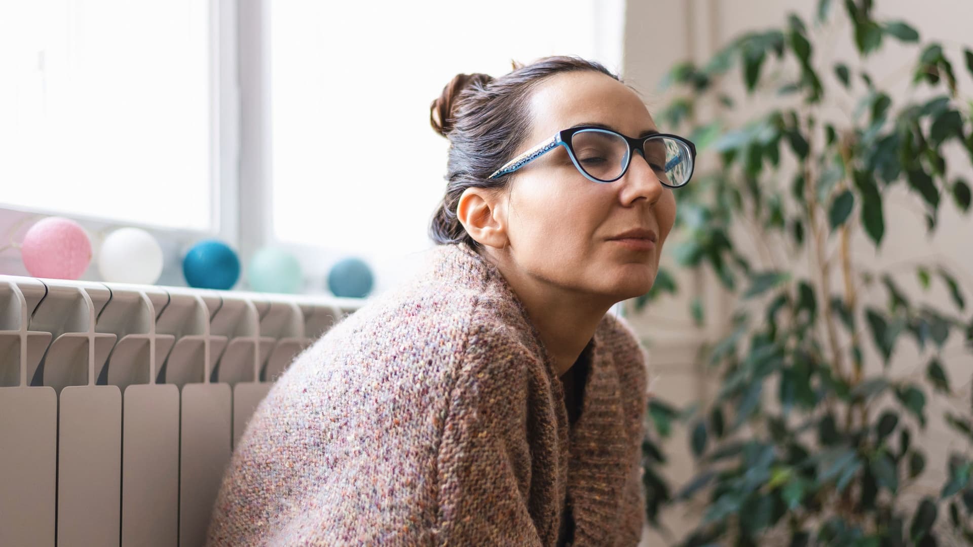 Eine Frau in einem Wollpulli lehnt mit geschlossenen Augen an einem Heizkörper