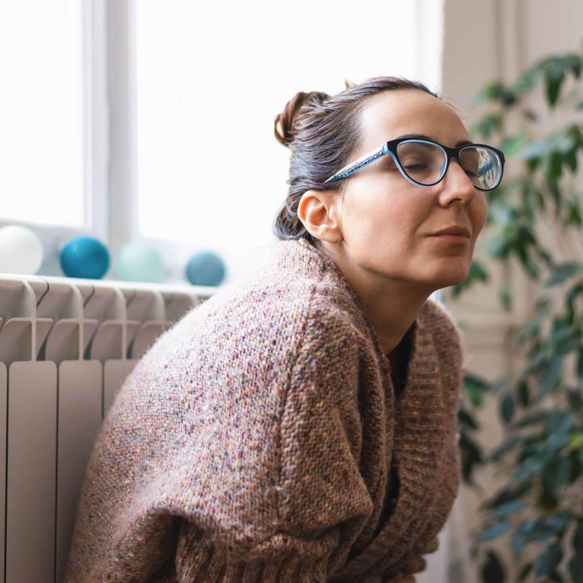Eine Frau in einem Wollpulli lehnt mit geschlossenen Augen an einem Heizkörper