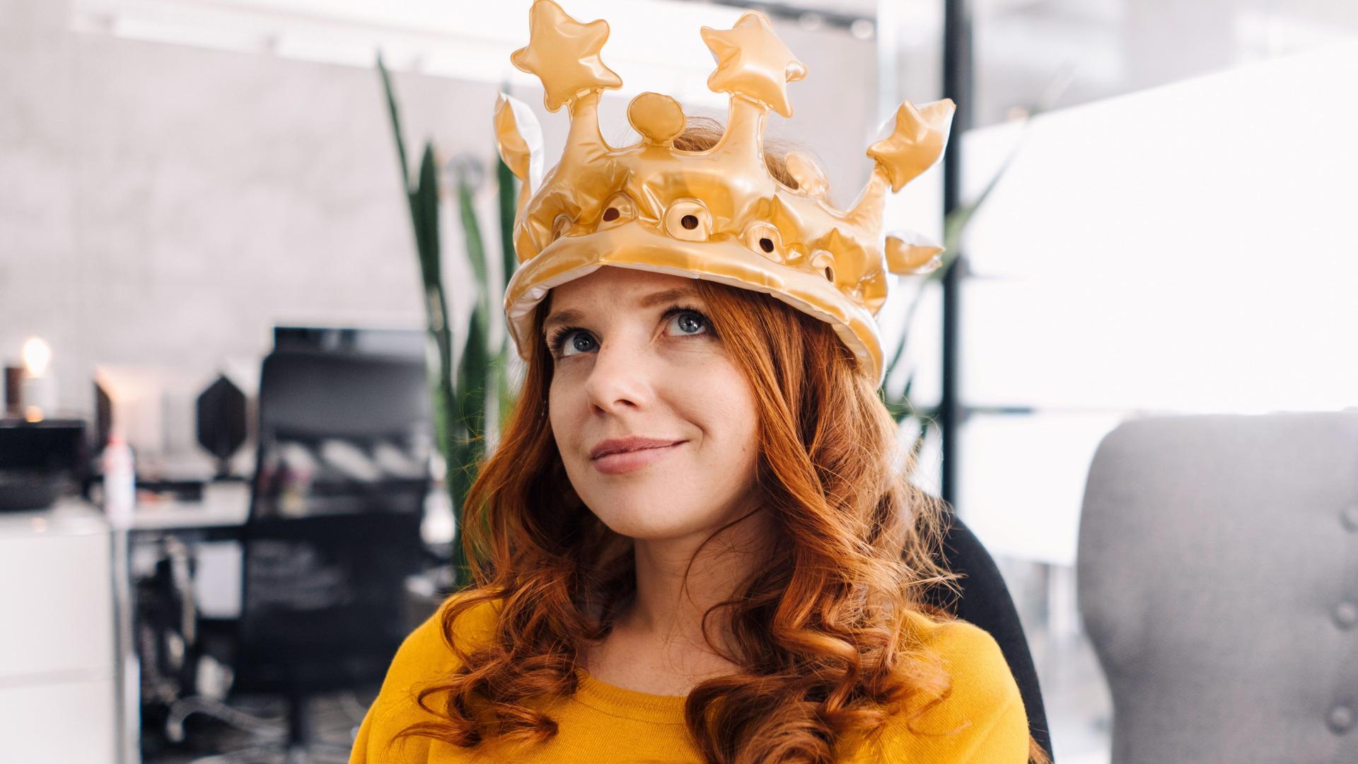 Junge Frau mit einer aufblasbaren Krone auf dem Kopf sitzt in einem Büro und blickt nach oben.