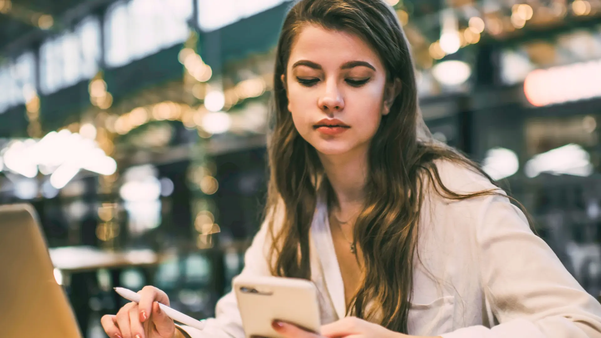 2. Zwei-Faktor-Authentifizierung Junge Frau vor Laptop schaut auf ihr Handy. Sie  sitzt in einer Markthalle an einem Tisch,