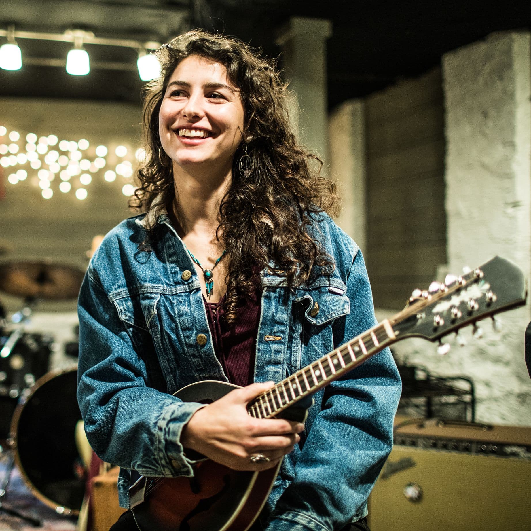 Eine junge Frau mit lockigen Haaren und Jeansjacke hält eine Mandoline und lächelt. Sie steht in einem Proberaum mit Verstärkern, Schlagzeug und Lichterkette im Hintergrund.