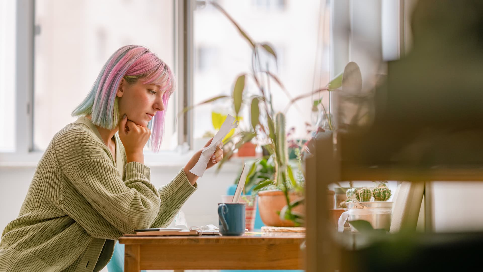 Frau mit bunten Haaren sitzt in einer Wohnung an einem Tisch und liest einen Brief. Sie hat den Kopf nachdenklich auf die geballte Hand gestützt.