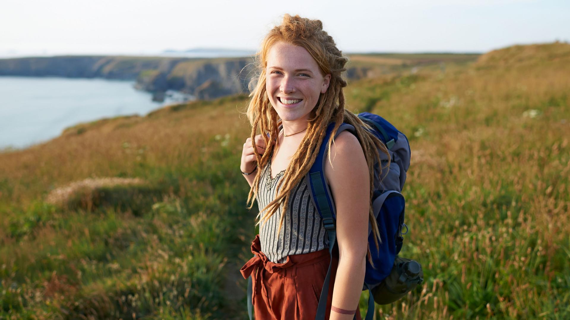 Eine junge Frau steht mit ihrem Rucksack auf einer Wiese an der Küste. Sie lächelt in die Kamera.