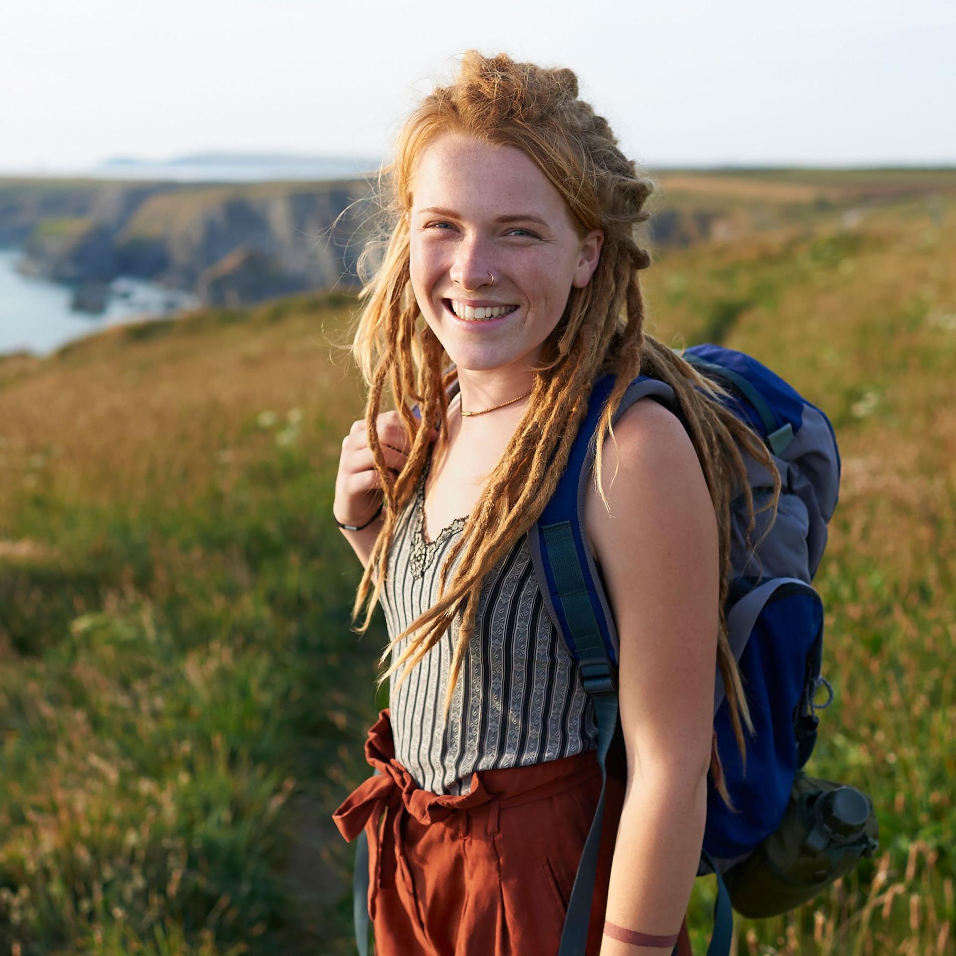 Eine junge Frau steht mit ihrem Rucksack auf einer Wiese an der Küste. Sie lächelt in die Kamera.