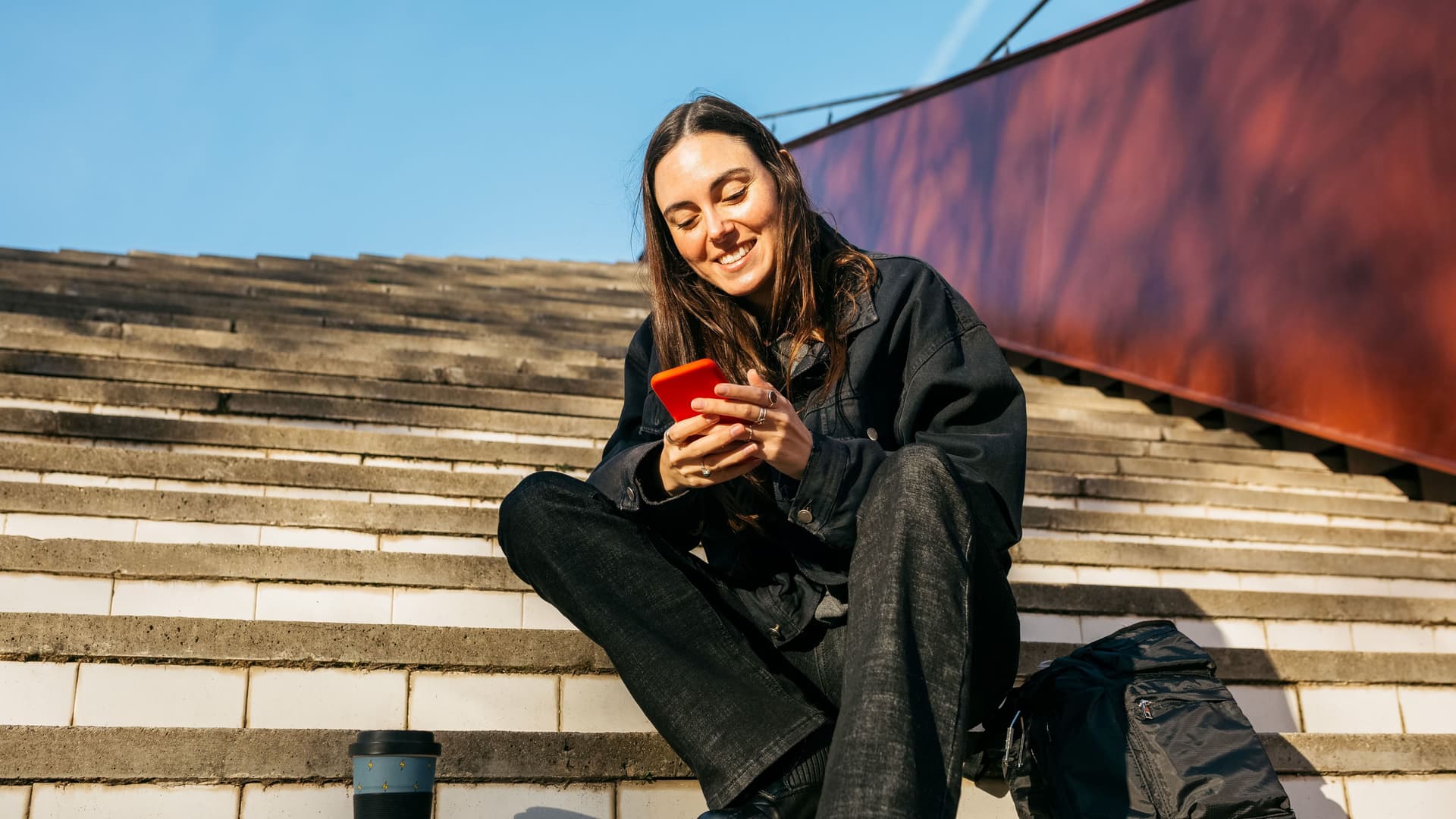 Junge Frau sitzt auf einer Treppe in der Sonne und schaut lächelnd auf ihr Smartphone.