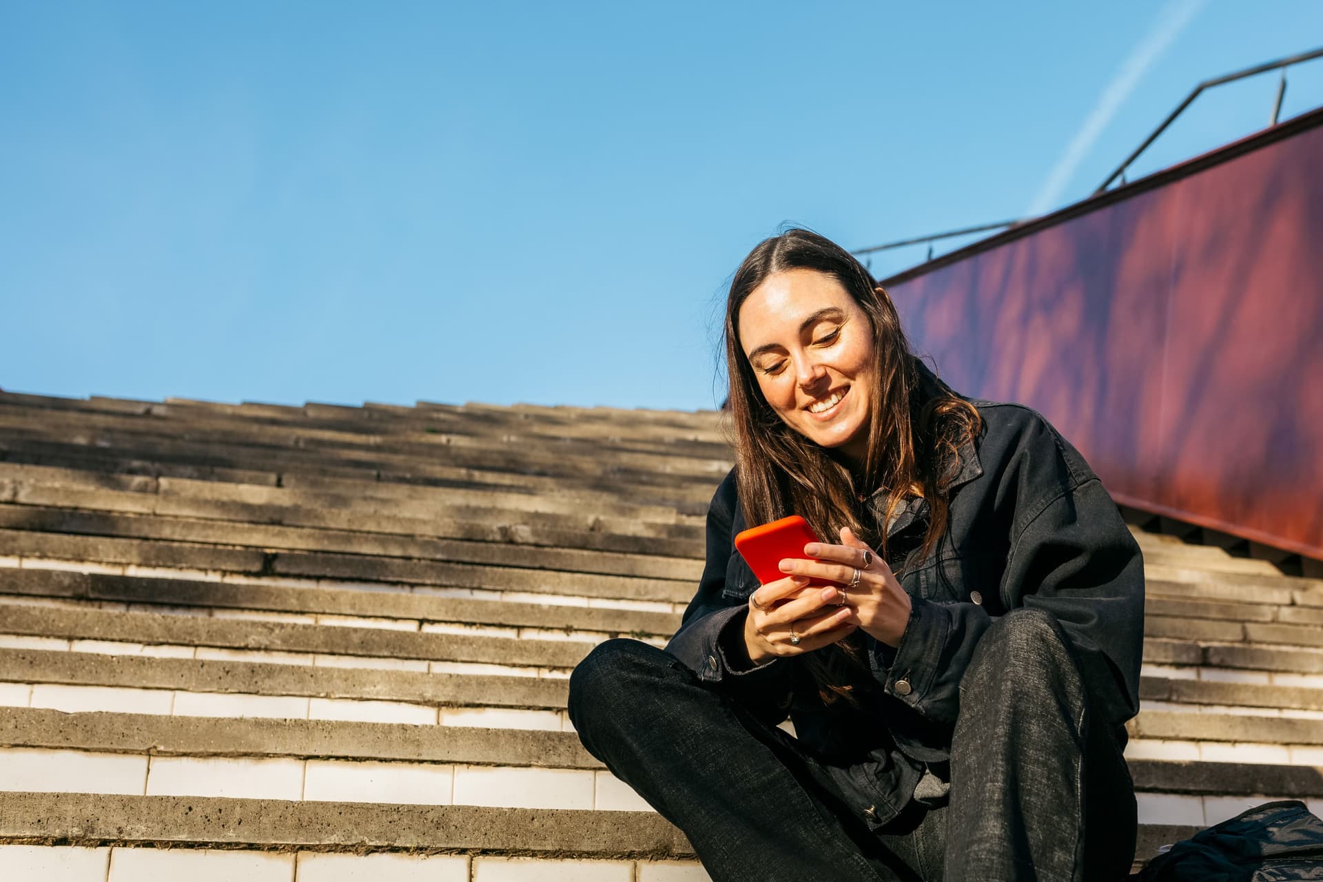 Junge Frau sitzt auf einer Treppe in der Sonne und schaut lächelnd auf ihr Smartphone.