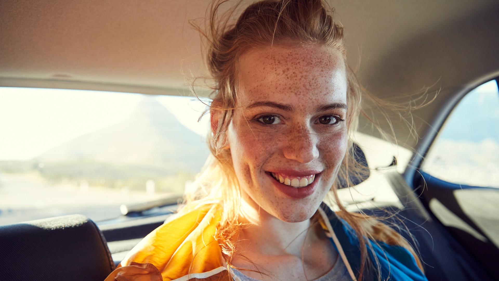 Junge Frau mit Sommersprossen sitzt auf dem Rücksitz eines Fahrzeuges. Sie lächelt.