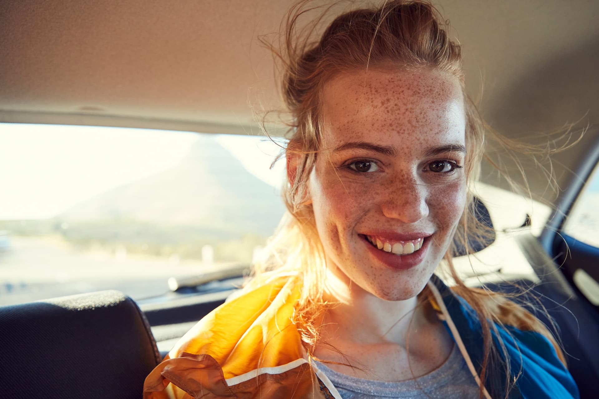 Junge Frau mit Sommersprossen sitzt auf dem Rücksitz eines Fahrzeuges. Sie lächelt.