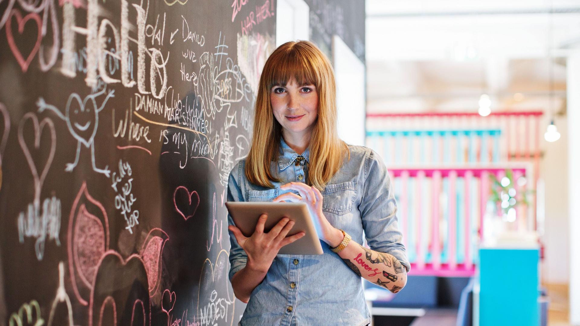 Junge Frau mit Tablet in der Hand steht in einem modernen Büro vor einer bunt bemalten Tafel und blickt in die Kamera.