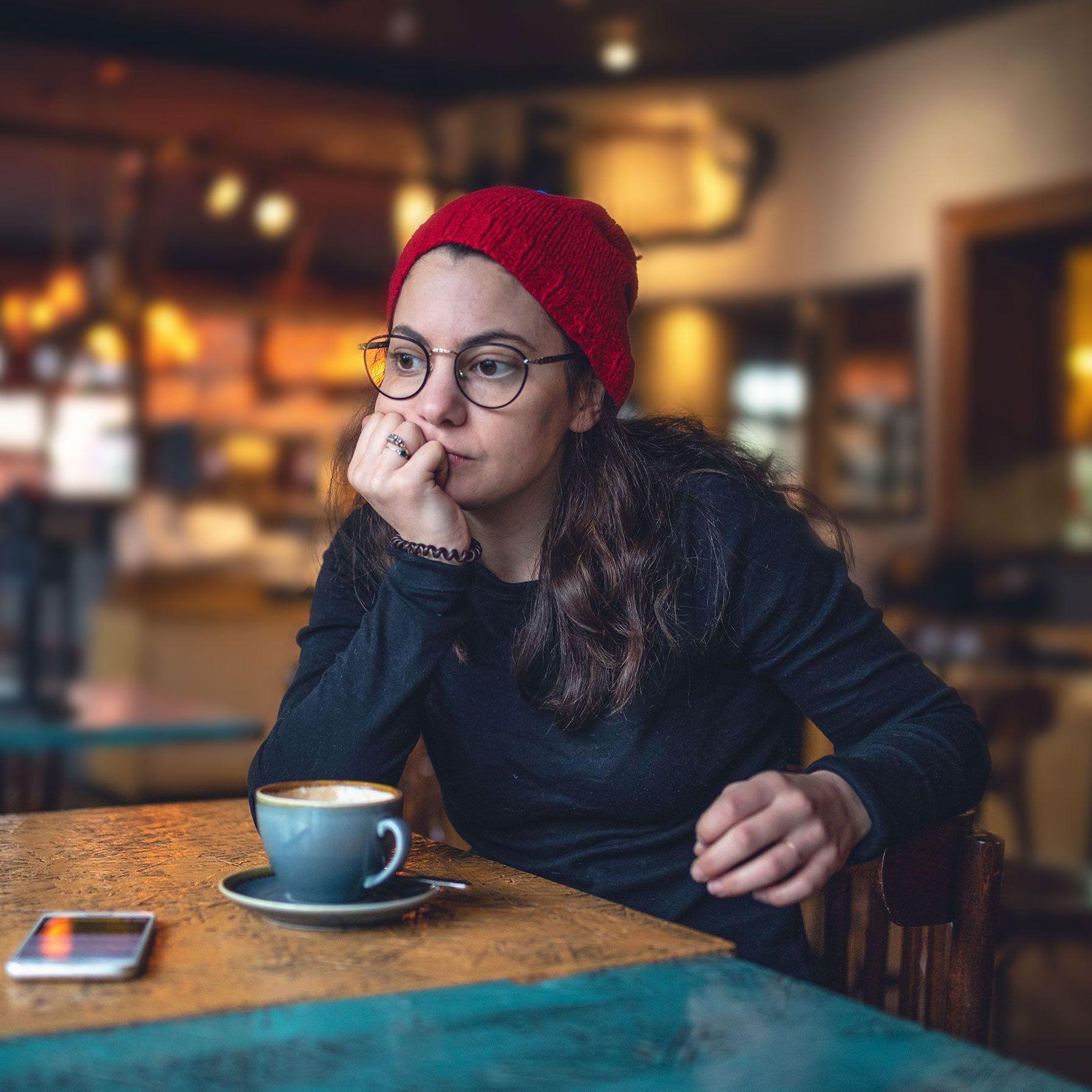 Eine junge Frau mit roter Mütze und runder Brille hat den Kopf auf die Hand gestützt und schaut ernst zum Cafe-Fenster. Vor ihr steht eine Kaffeetasse und ein Handy.