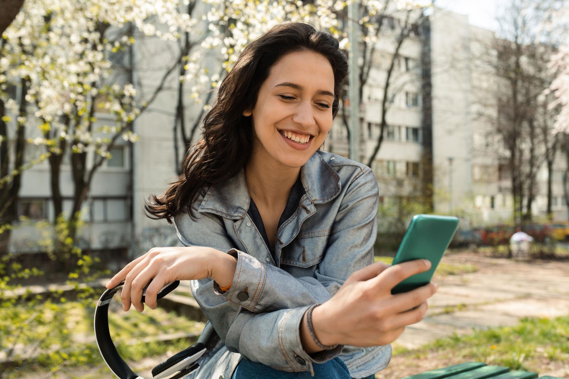 EIne junge schwarzhaarige Frau, auf ihrem Fahrrad schaut lächelnd auf ihr Smartphone. Im Hintergrund blühende Bäume und Sträucher.