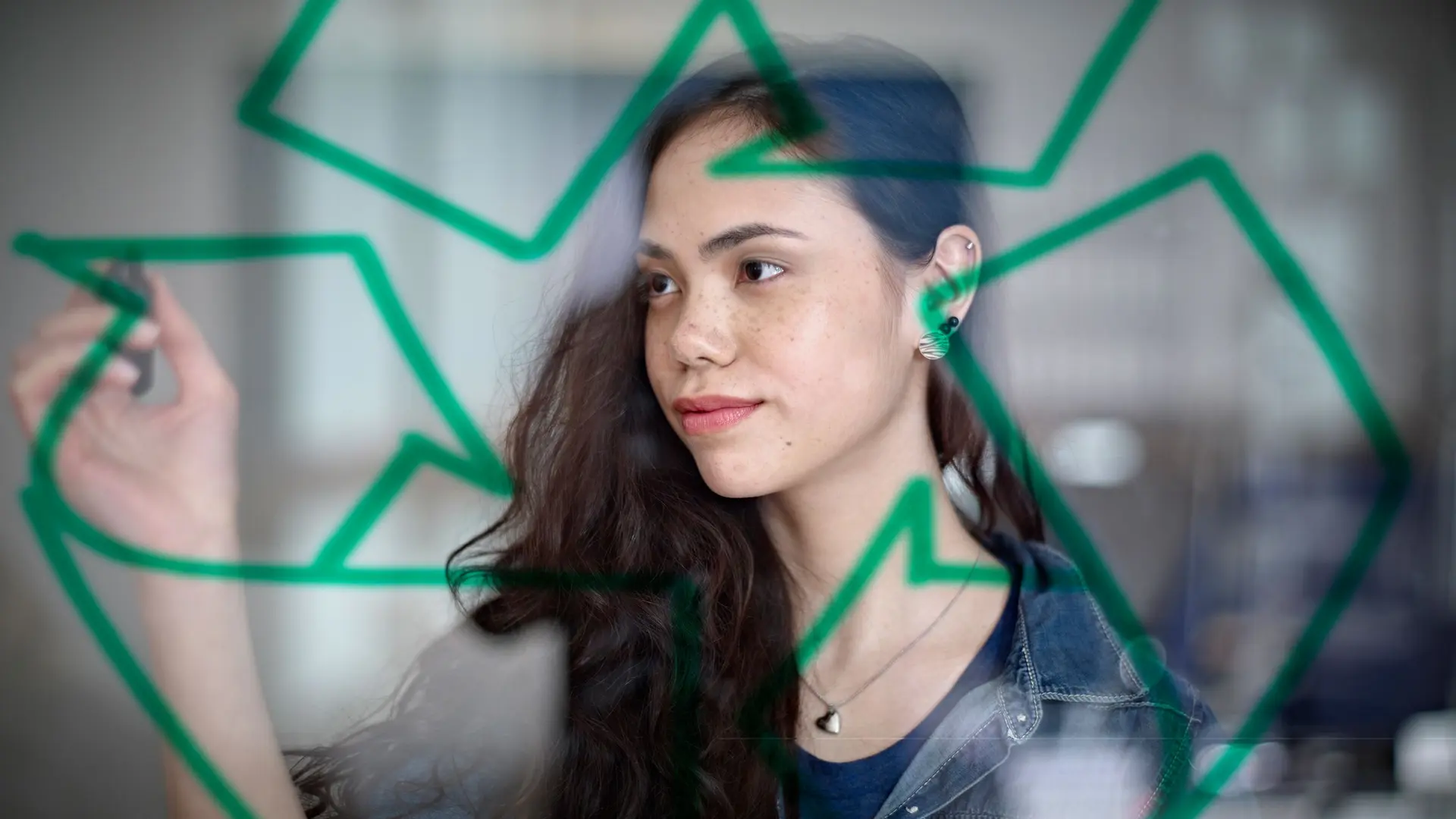 Eine junge Frau steht vor einer Glaswand und zeichnet mit einem Stift das Recycling-Symbol darauf. Sie blickt konzentriert und nachdenklich, während das Symbol im Vordergrund deutlich zu sehen ist.