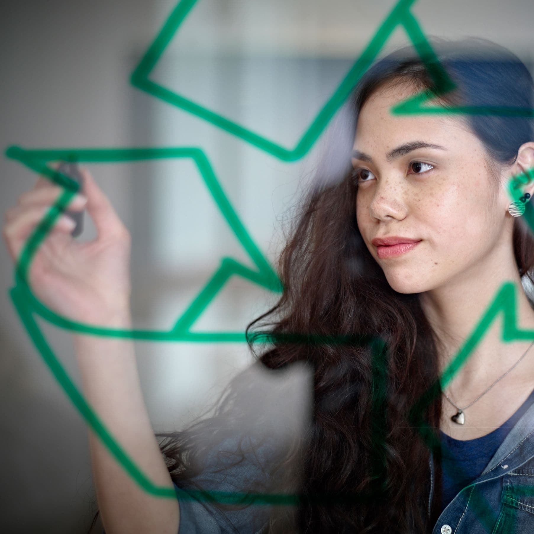 Eine junge Frau steht vor einer Glaswand und zeichnet mit einem Stift das Recycling-Symbol darauf. Sie blickt konzentriert und nachdenklich, während das Symbol im Vordergrund deutlich zu sehen ist.