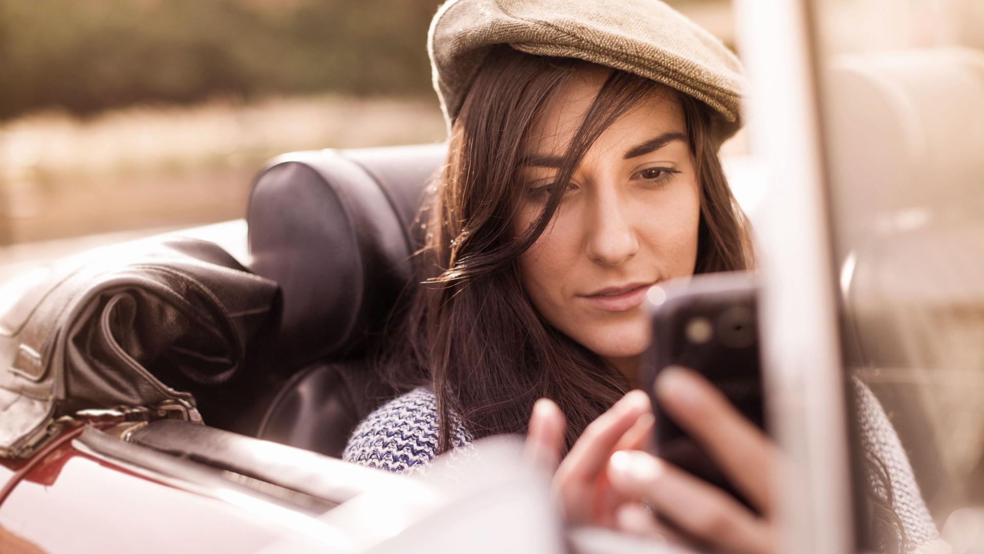 Junge Frau mit einer Schirmmütze sitzt in einem Cabriolet und verwendet ihr Smartphone