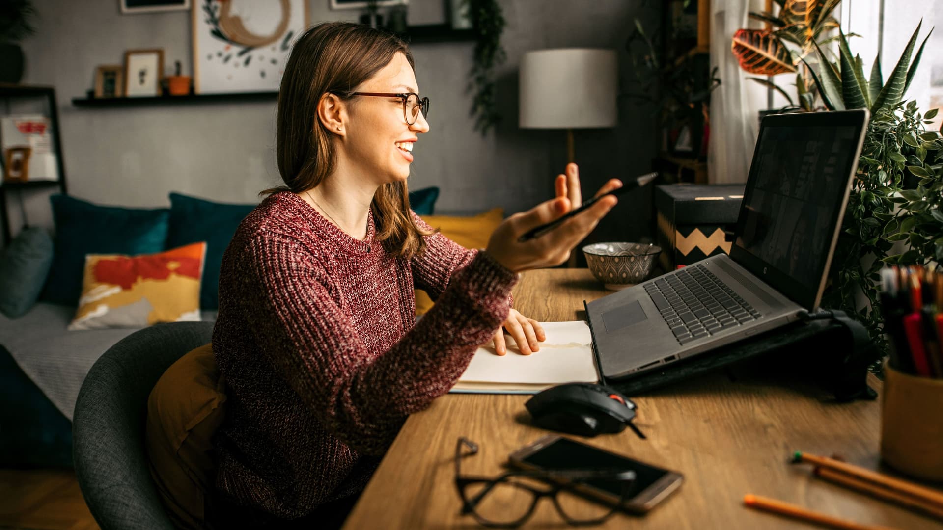 Junge Frau sitzt während einer Onlinekonferenz gestikulierend vor ihrem Laptop in ihrer Wohnung.