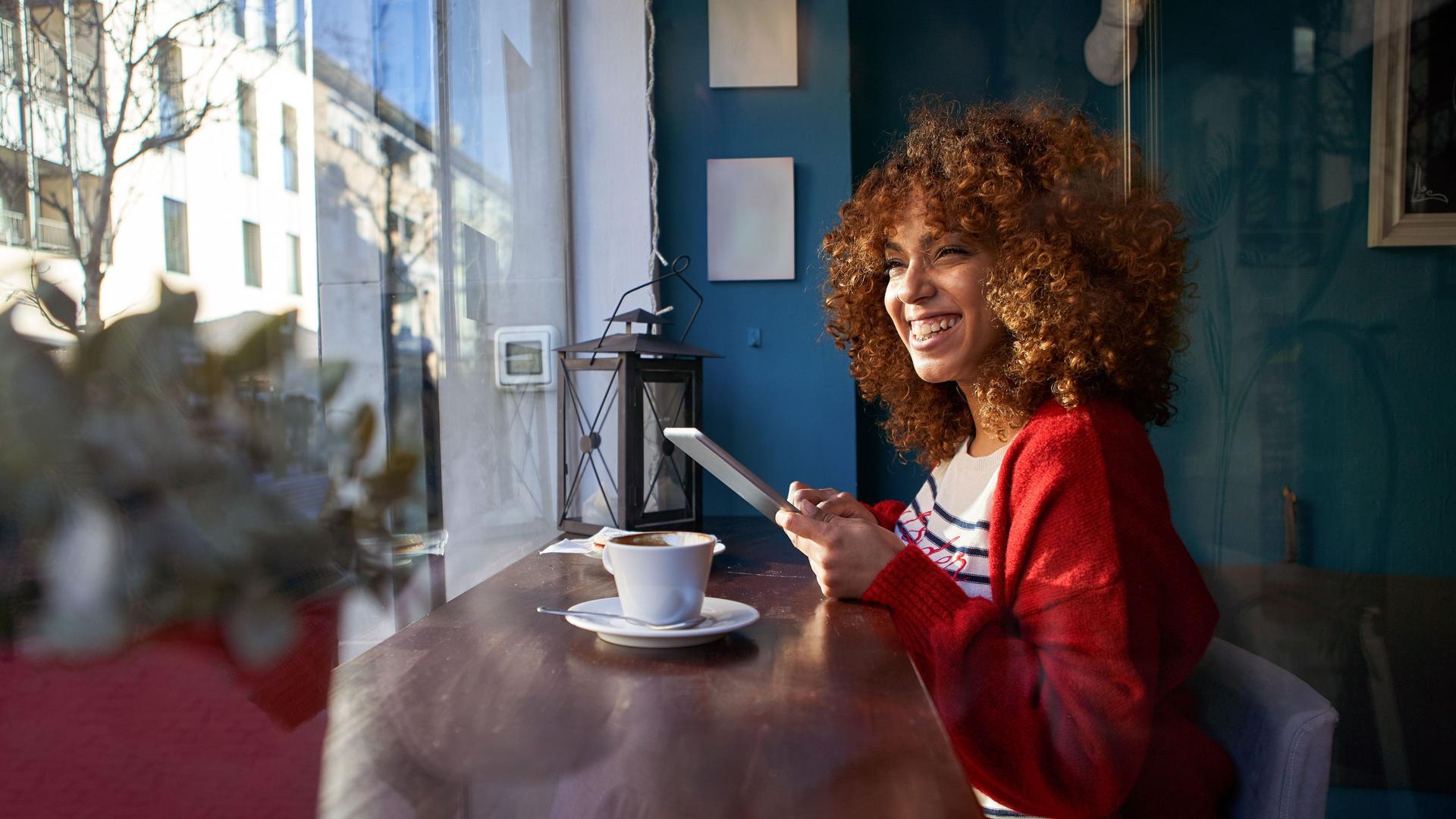 Eine junge Frau sitzt lachend im Cafe, blickt aus dem Fenster und hält ein Mobiltelefon in der Hand