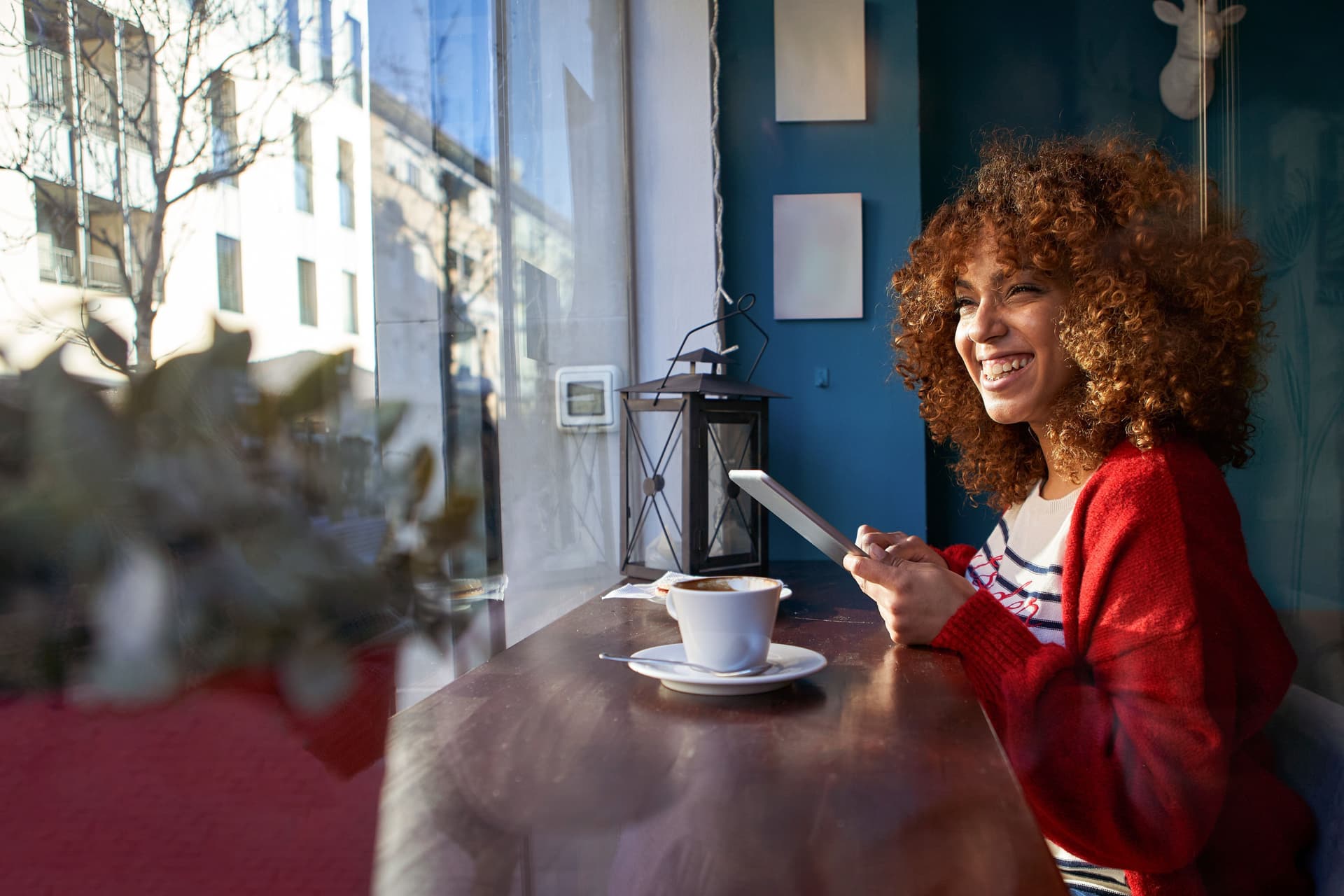 Eine junge Frau sitzt lachend im Cafe, blickt aus dem Fenster und hält ein Mobiltelefon in der Hand