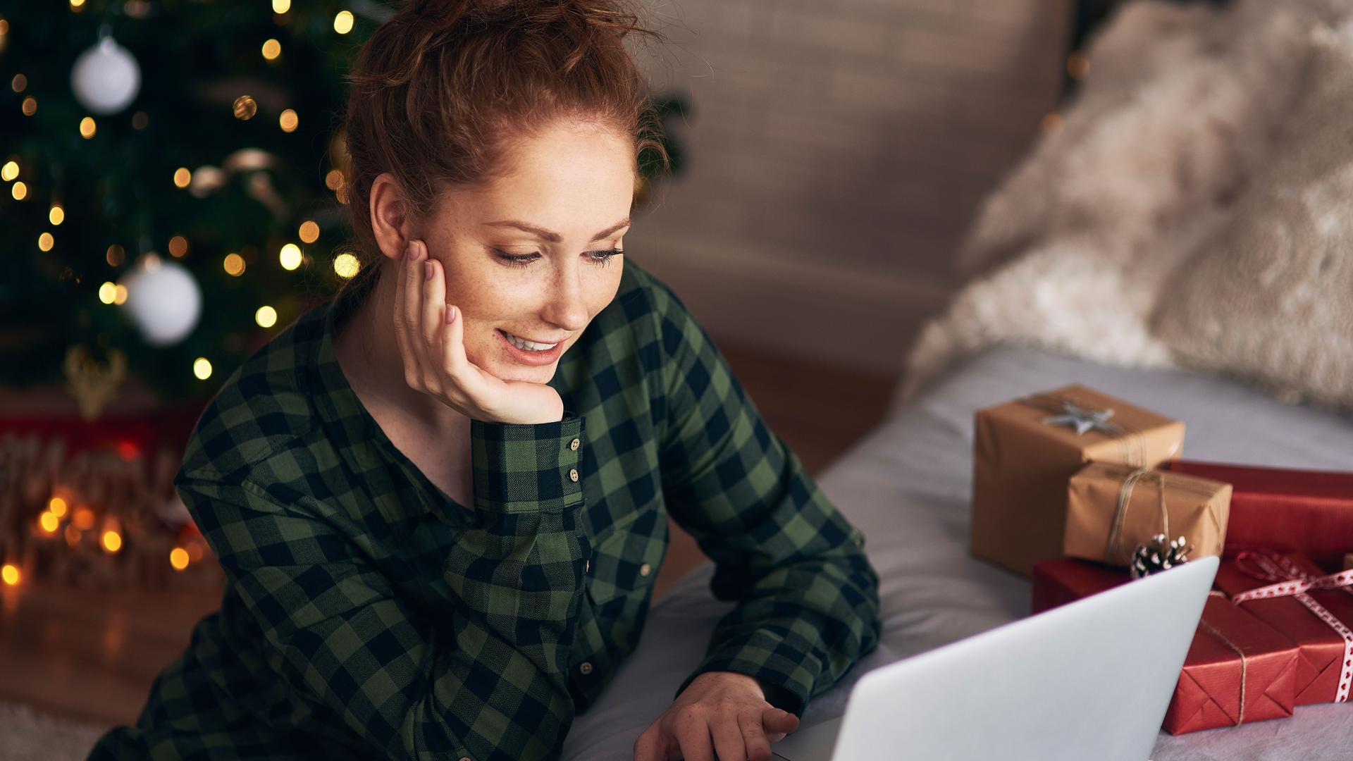 Eine junge Frau arbeitet vor dem Weihnachtsbaum am Boden sitzend an einem Laptop, der auf einem Sofa steht.