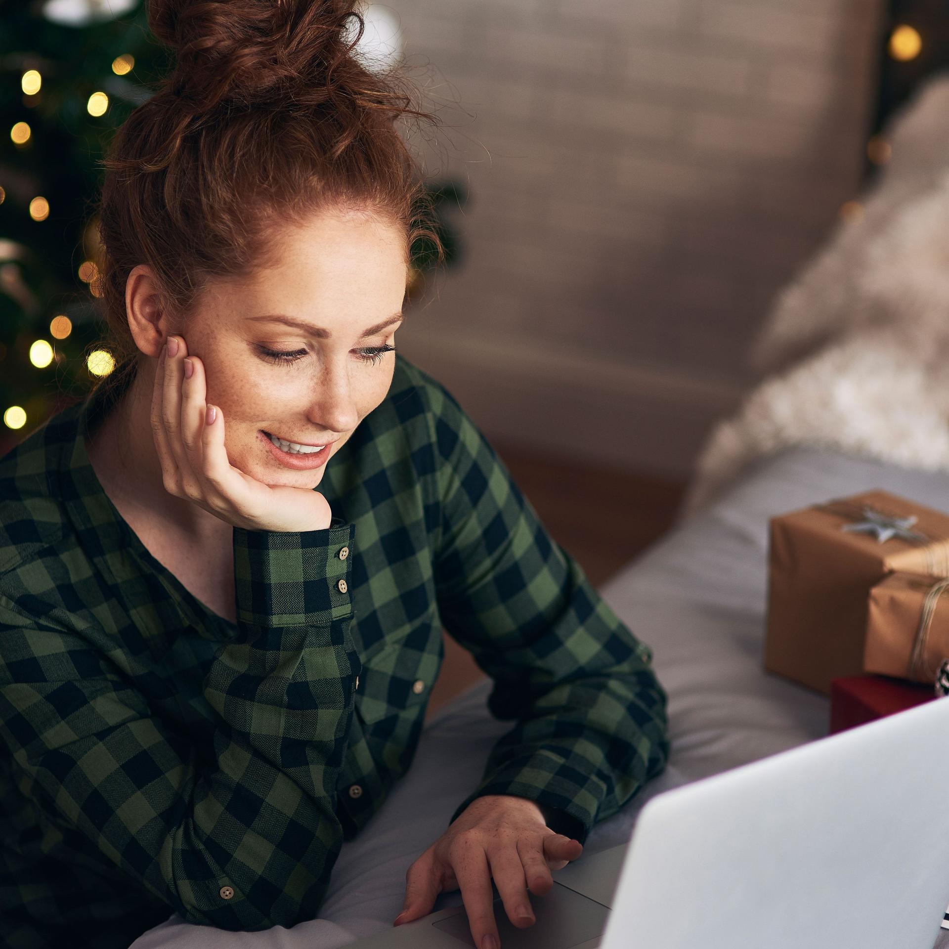 Eine junge Frau arbeitet vor dem Weihnachtsbaum am Boden sitzend an einem Laptop, der auf einem Sofa steht.