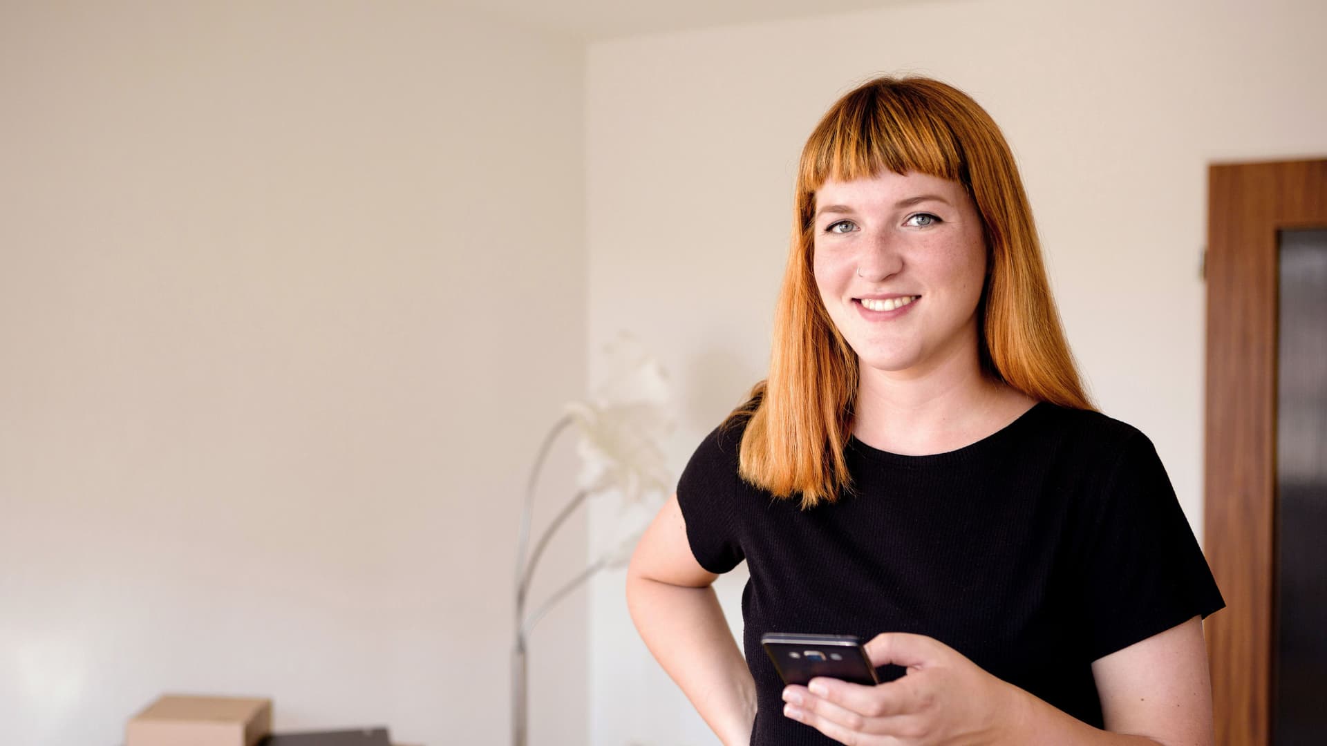 Junge Frau blickt während des Umzugs lächelnd in die Kamera und hält ein Smartphone in der Hand