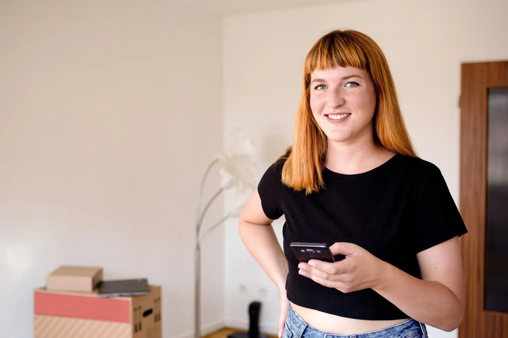 Junge Frau blickt während des Umzugs lächelnd in die Kamera und hält ein Smartphone in der Hand