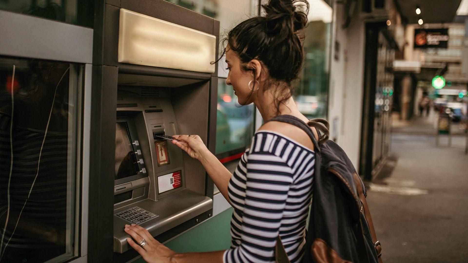 Eine junge Frau mit Dutt und Rucksack steht vor einem Straßen-Bankautomaten und führt ihre Bankkarte ein. 