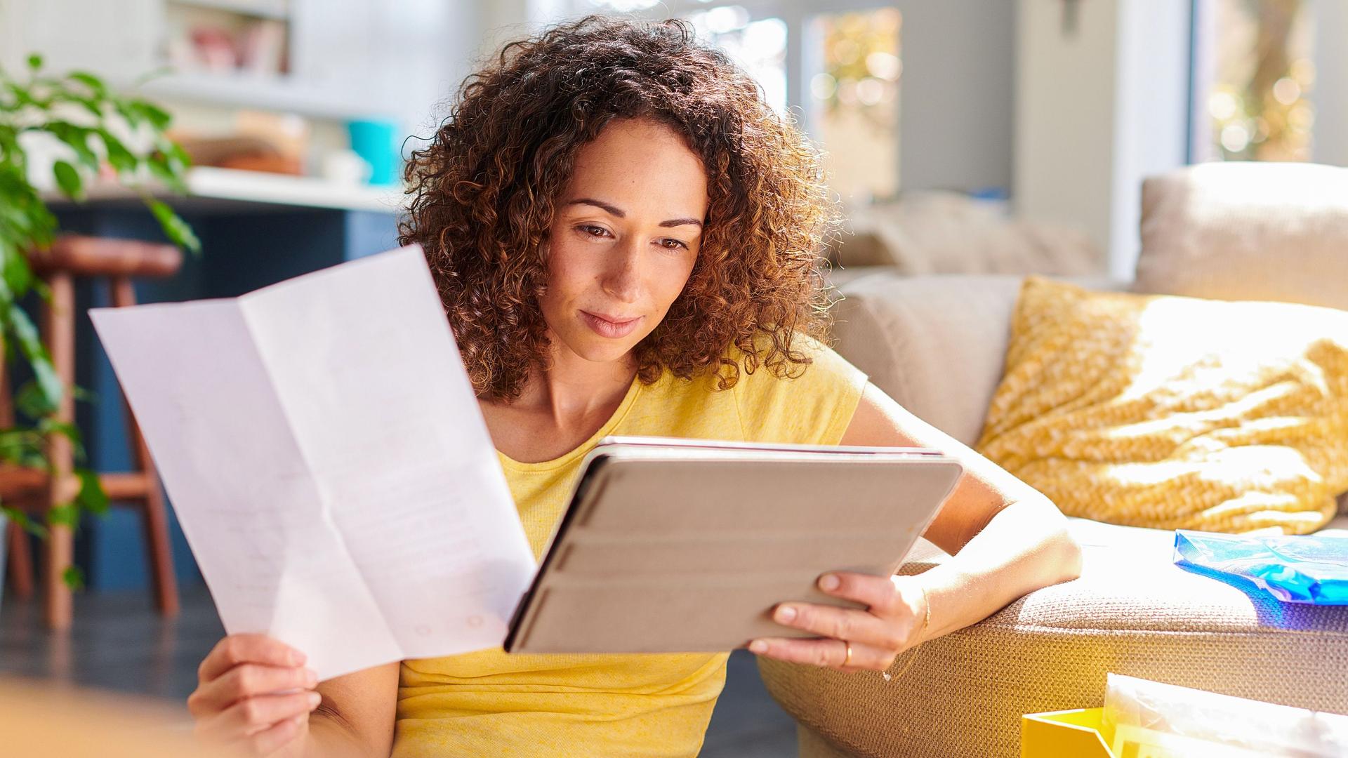 Eine junge Frau sitzt in einem hellen Wohnzimmer vor dem Sofa auf dem Boden und schaut auf ein Tablet in ihrer linken Hand. In der anderen Hand hält sie ein Stück Papier.