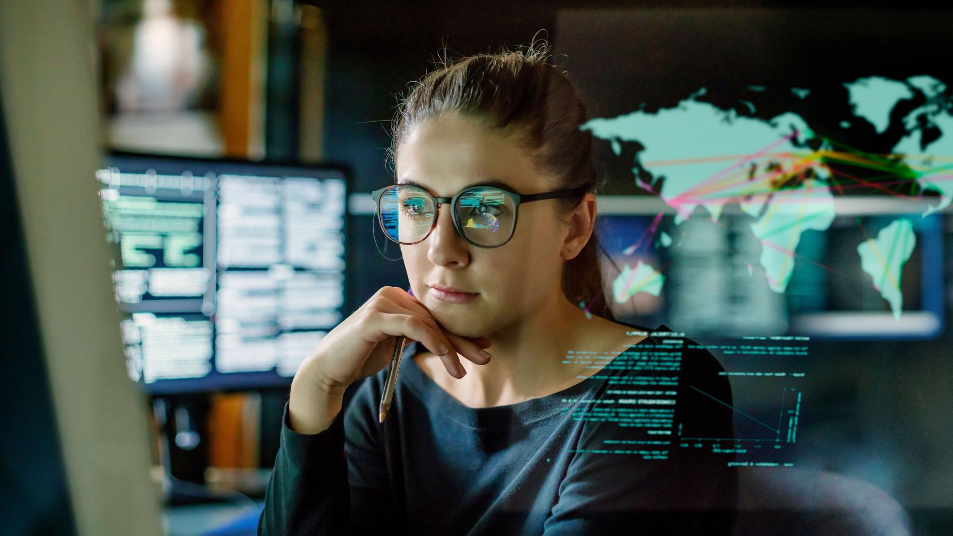 Jungen Frau mit Brille, die in einem dunklen Büro von Computermonitoren umgeben ist. Vor ihr befindet sich ein durchsichtiges Display, das eine Weltkarte mit einigen Daten zeigt.