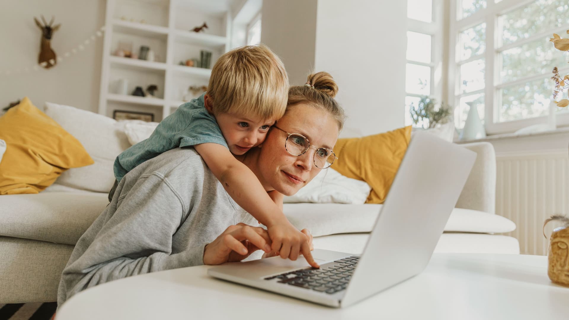 Ein junge Frau und ein kleines Kind sitzen vor einem Sofa und tippen auf Laptop.