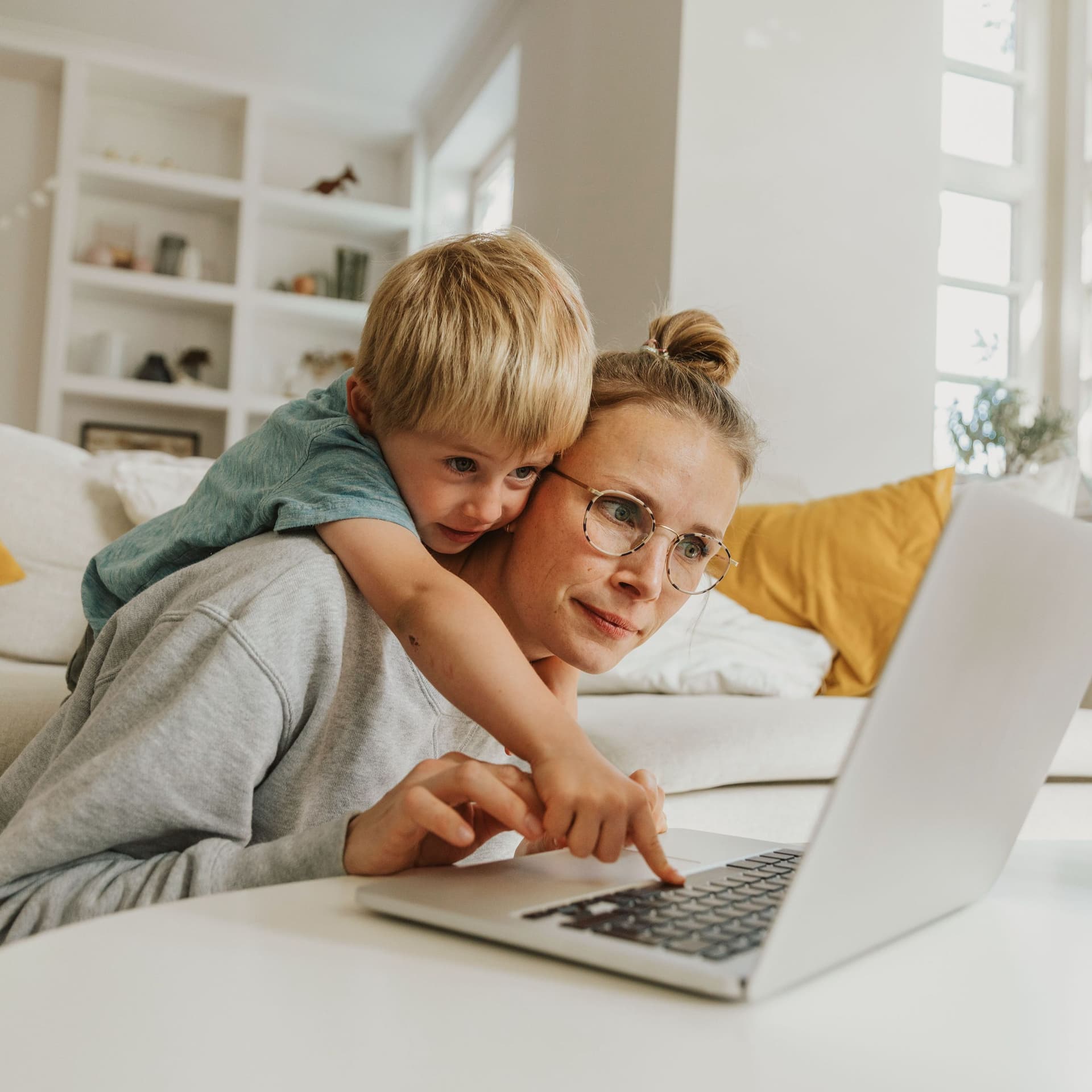 Ein junge Frau und ein kleines Kind sitzen vor einem Sofa und tippen auf Laptop.