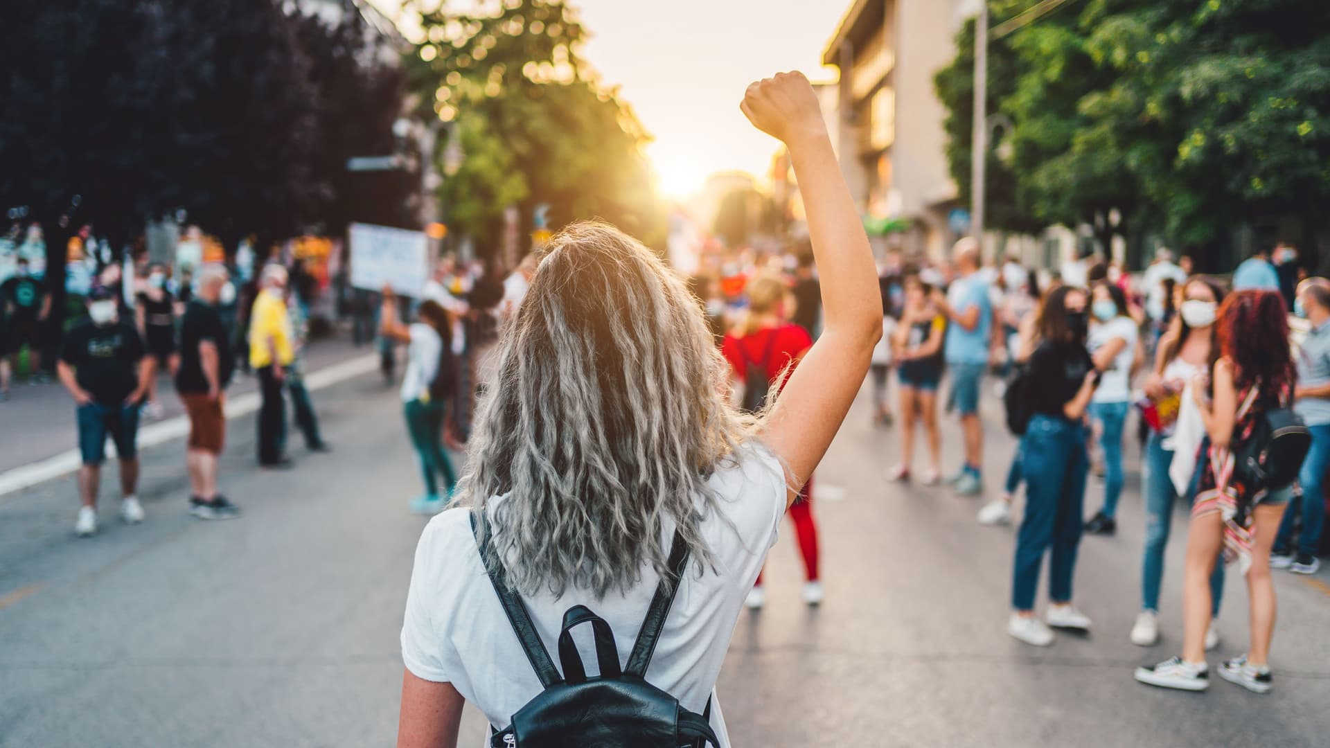 Junge Frau mit erhobener Faust, die auf der Straße protestiert. Sie steht mit dem Rücken zur Kamera, im Hintergrund andere Demonstrierende und ein Sonnenuntergang.