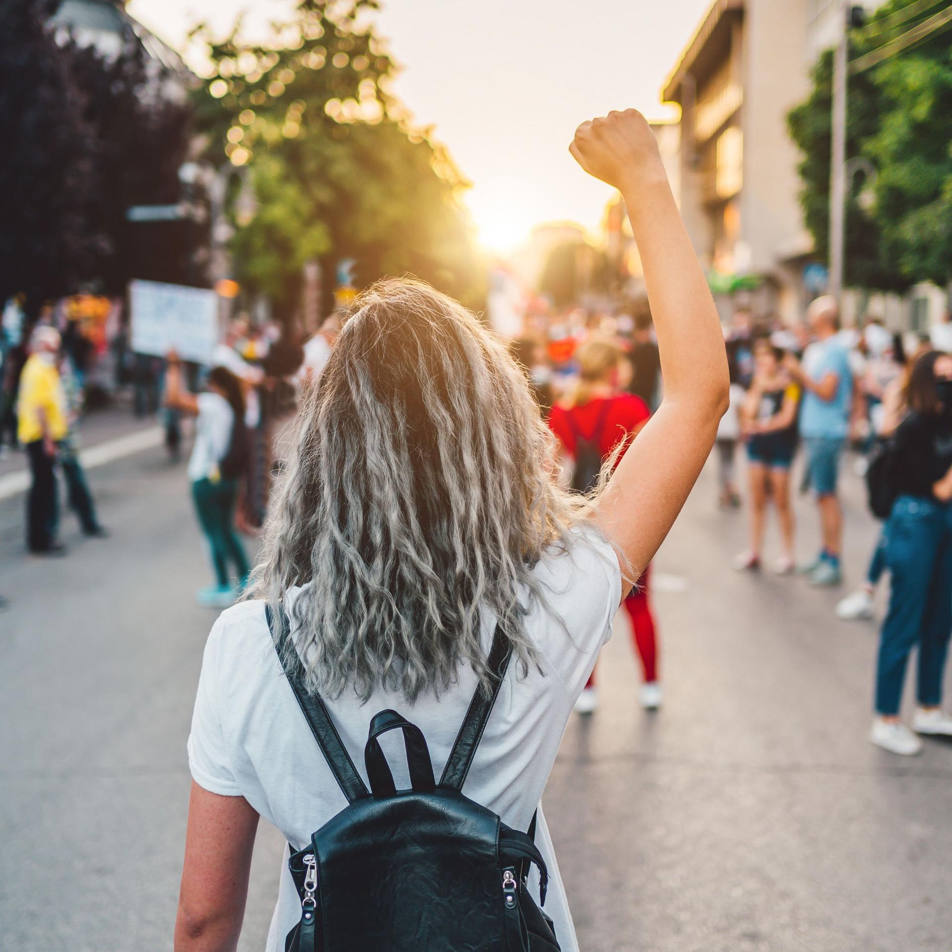 Junge Frau mit erhobener Faust, die auf der Straße protestiert. Sie steht mit dem Rücken zur Kamera, im Hintergrund andere Demonstrierende und ein Sonnenuntergang.