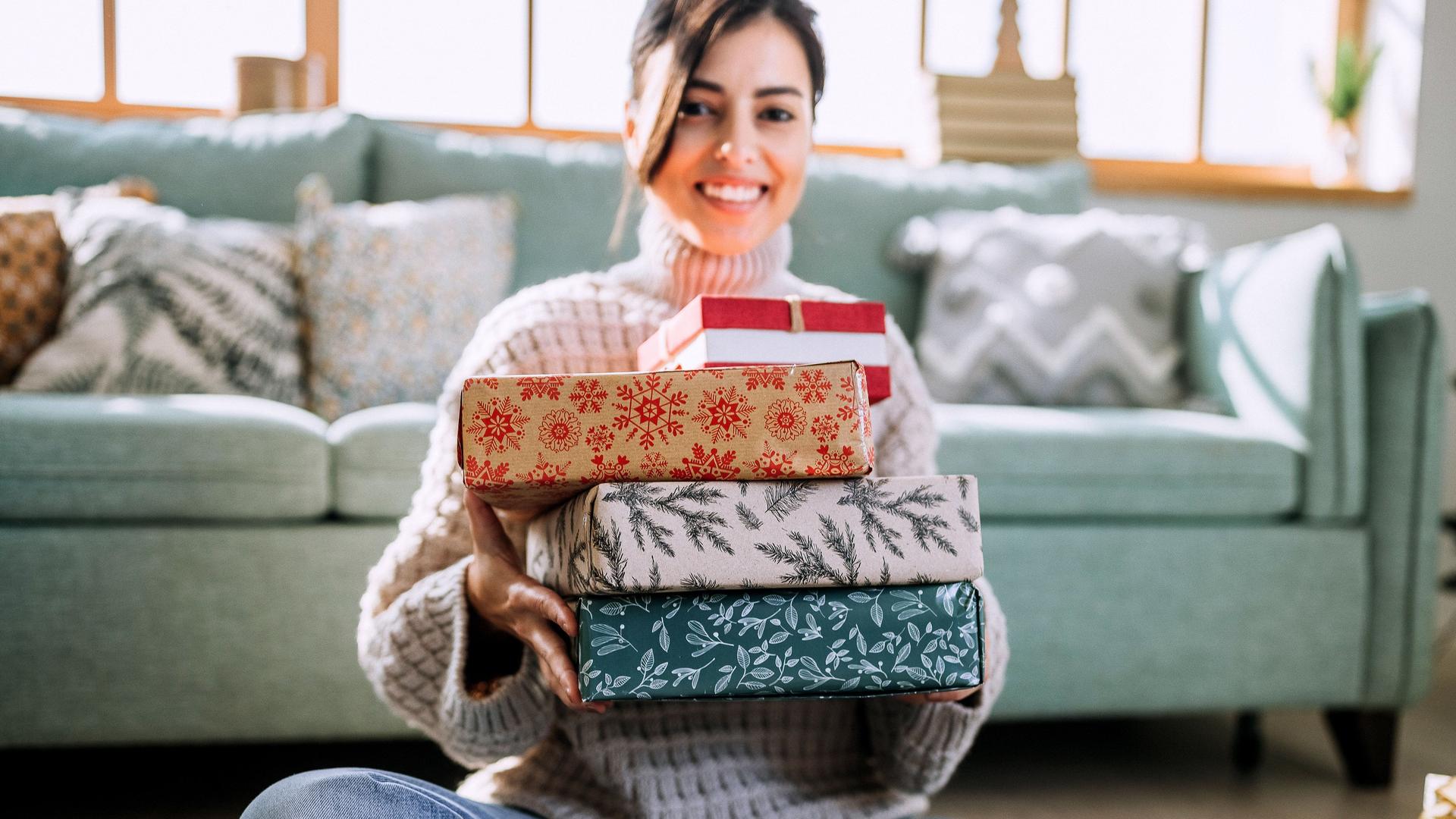 Porträt einer jungen Frau, die Weihnachtsgeschenke präsentiert während sie im Wohnzimmer vor der Couch auf den Boden sitzt