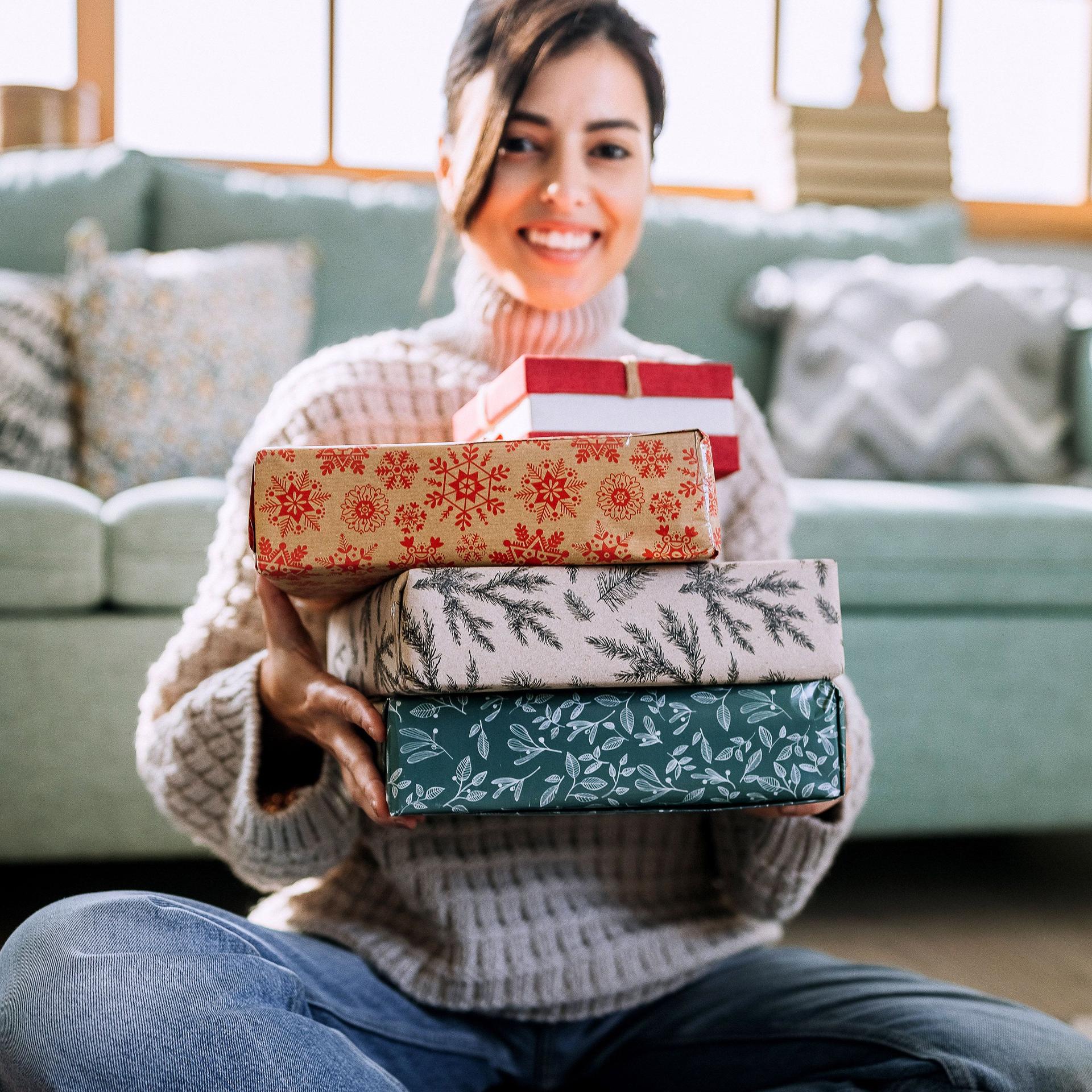 Porträt einer jungen Frau, die Weihnachtsgeschenke präsentiert während sie im Wohnzimmer vor der Couch auf den Boden sitzt