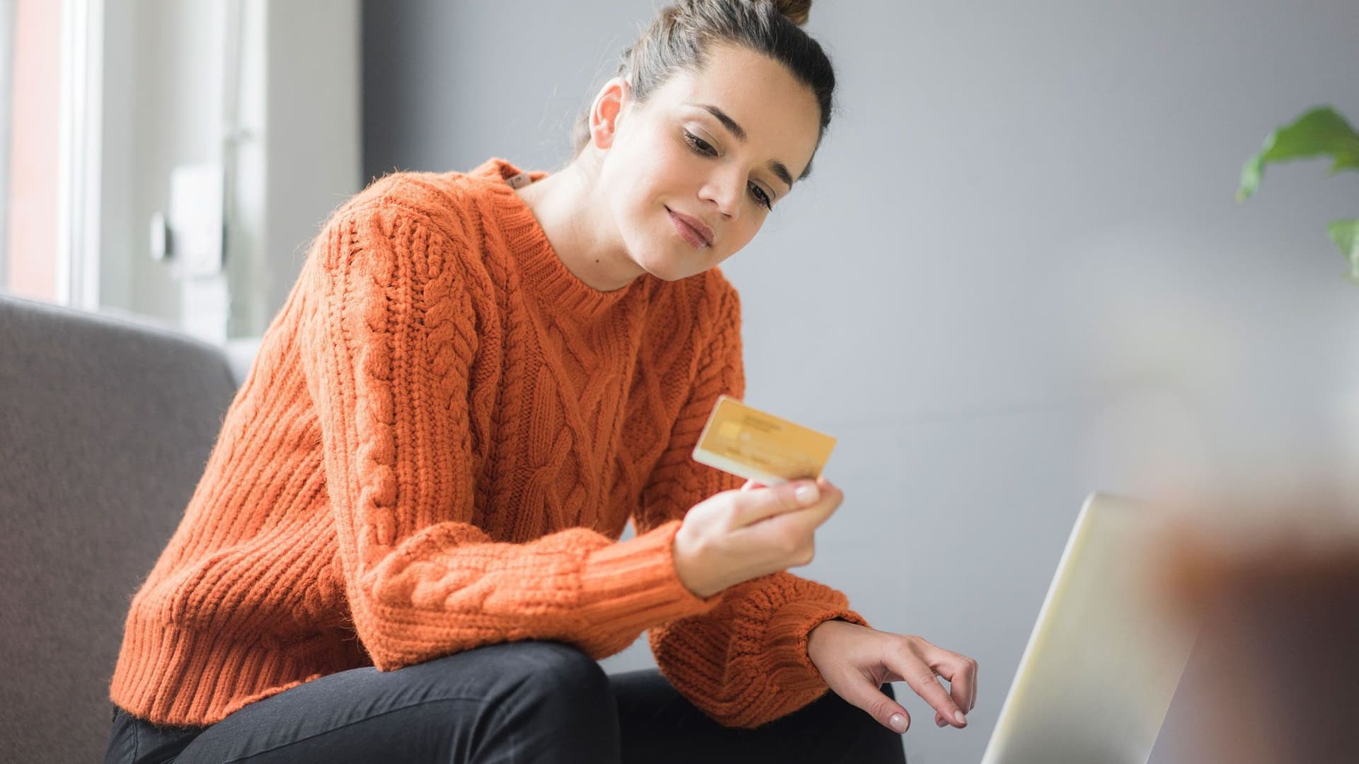 Junge Frau mit einer Bankkarte in der Hand sitzt auf einem Sessel und betrachtet die Rückseite der Karte. Vor ihr ist ein Laptop aufgebaut.