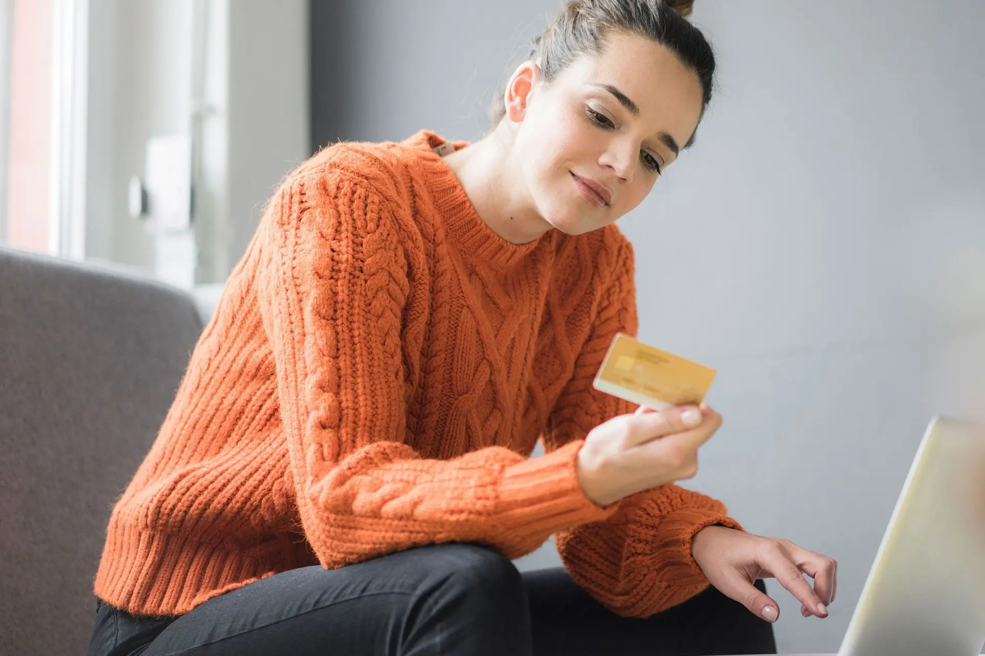 Junge Frau mit einer Bankkarte in der Hand sitzt auf einem Sessel und betrachtet die Rückseite der Karte. Vor ihr ist ein Laptop aufgebaut.