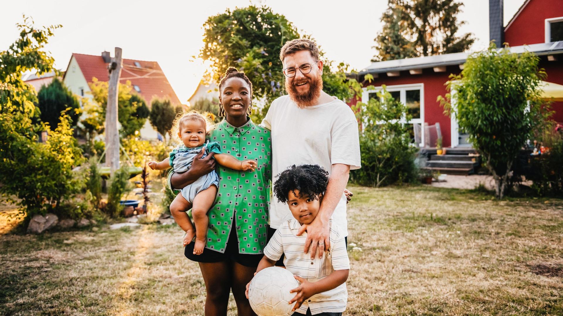 Eine Familie mit einem kleinen Kind und einem Baby steht vor einem roten Einfamilienhaus.