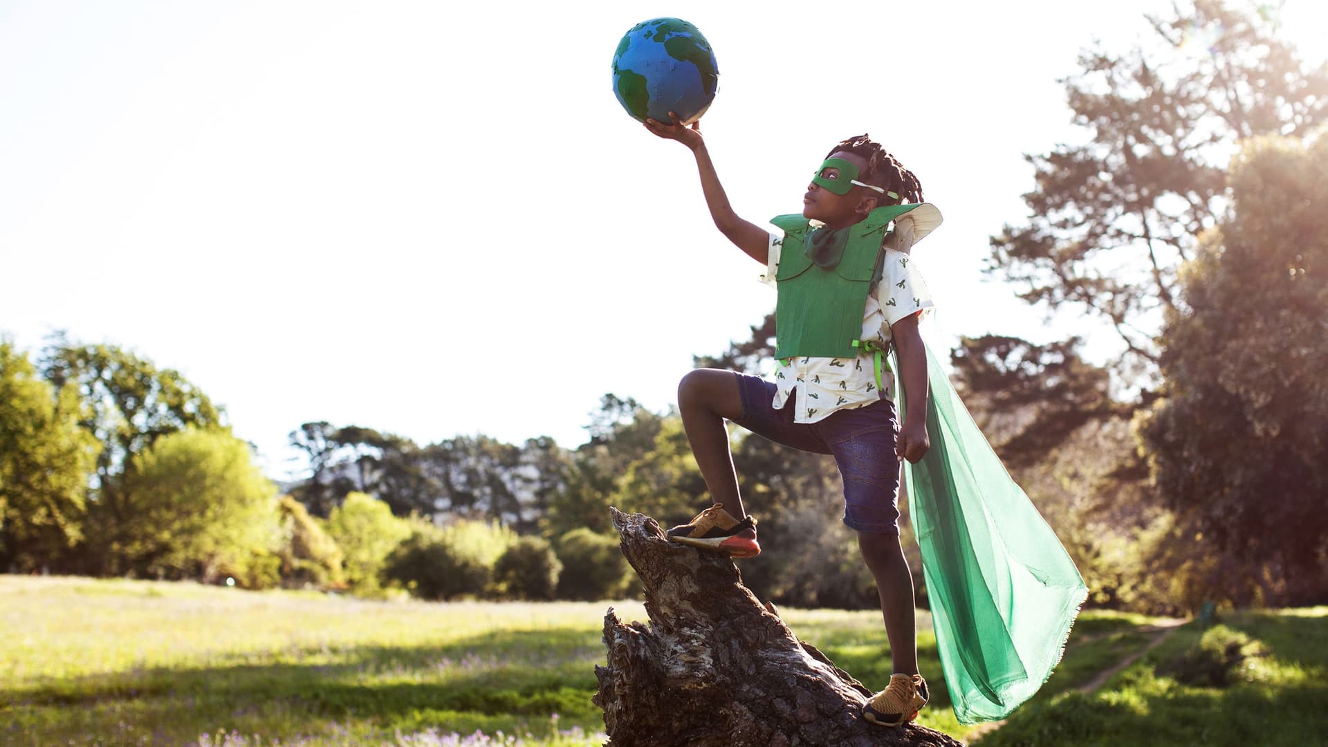 Kind mit grünem Superheldenkostüm steht auf Baumstamm und hält eine Erdkugel in die Luft in sonniger Parklandschaft